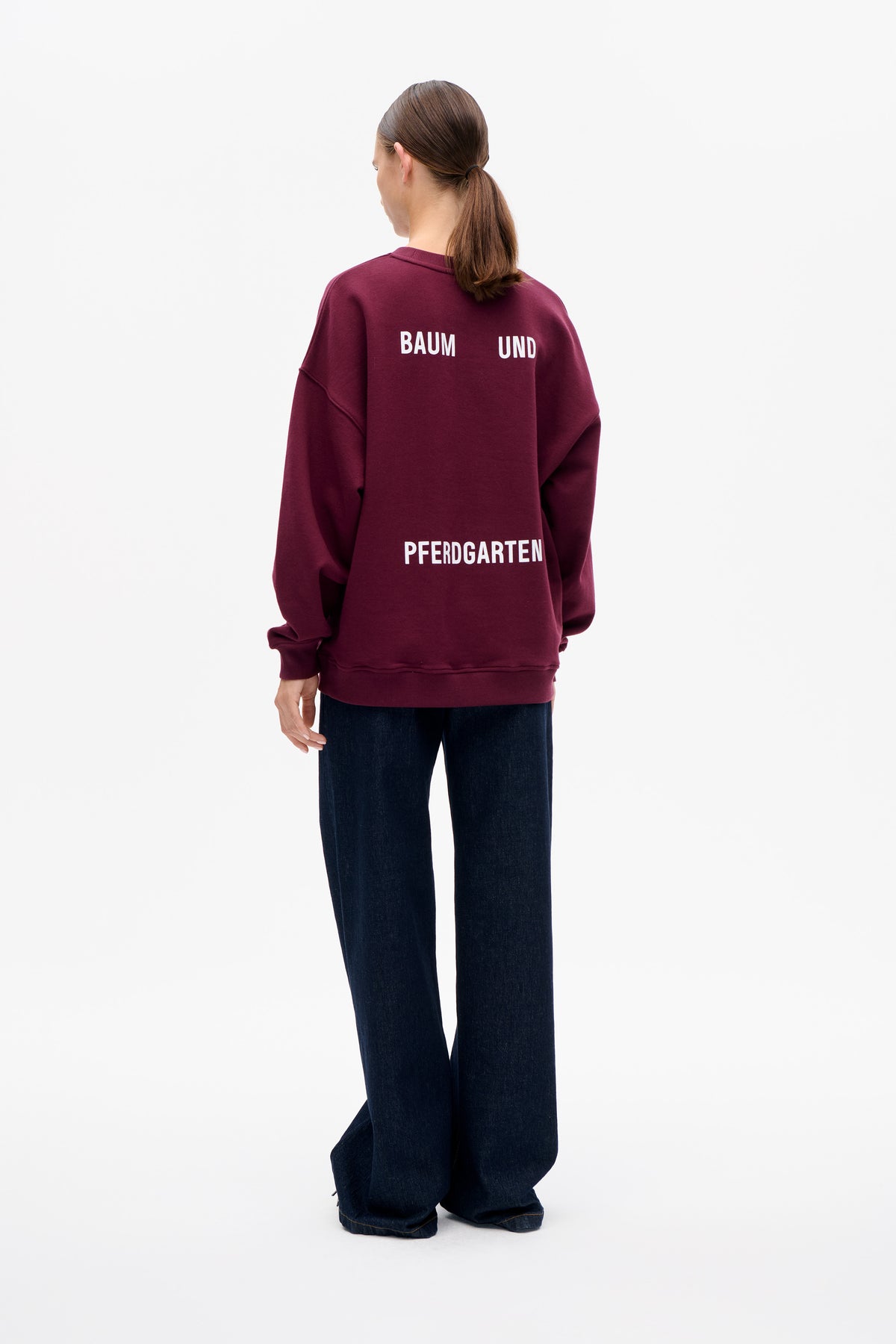 Achteraanzicht van een model in een bordeauxrode oversized sweatshirt met witte Baum und Pferdgarten-opdruk en donkere jeans met wijde pijpen.