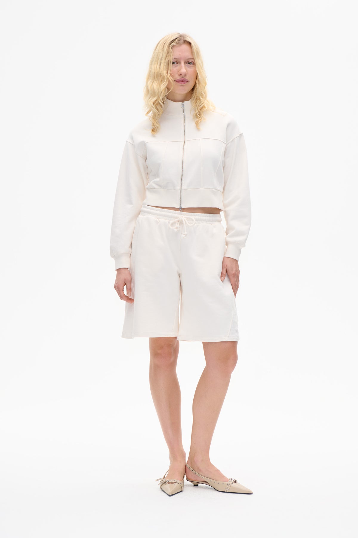 Modèle portant une veste ivoire zippée à l'avant avec coutures en panneaux, un short assorti à cordon et des ballerines pointues beige sur fond blanc.