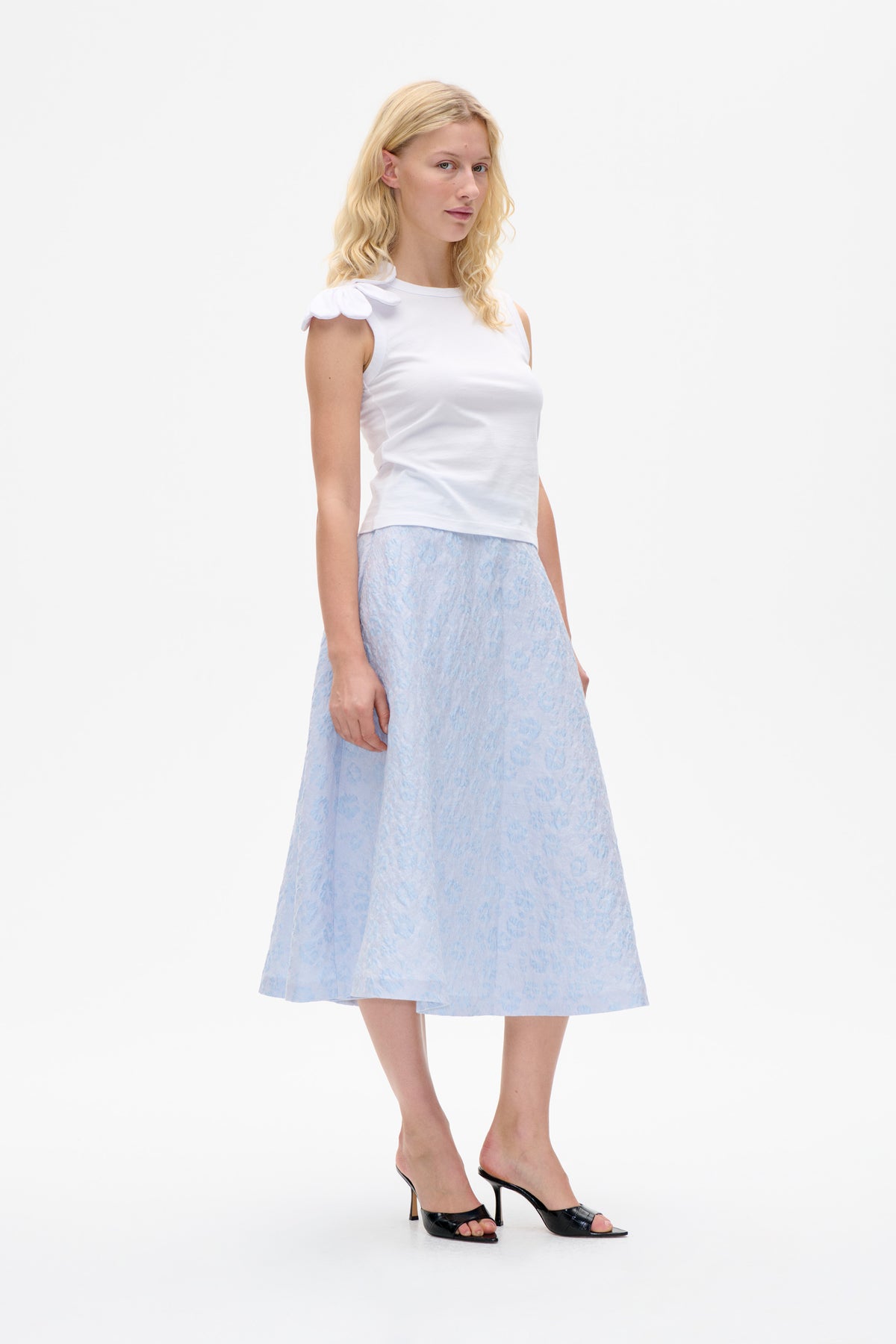 Le mannequin porte un haut sans manches blanc, orné d’un détail en forme de pétale aux épaules, et une jupe midi imprimée bleu pâle, sur un fond de studio blanc.