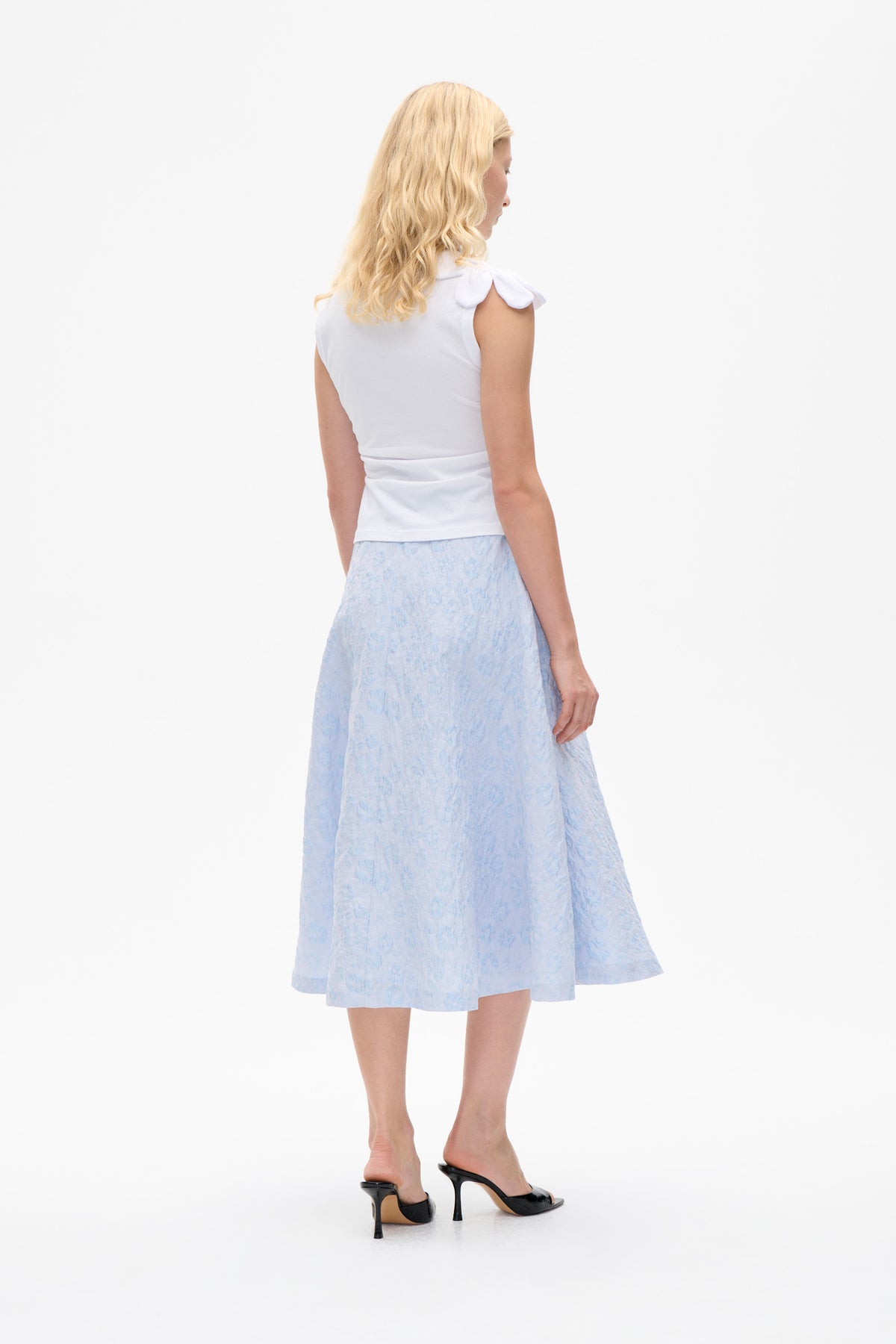 Mannequin présenté de dos portant un haut blanc sans manches avec des détails aux épaules en forme de nœud et une jupe midi à fleurs bleu pâle.