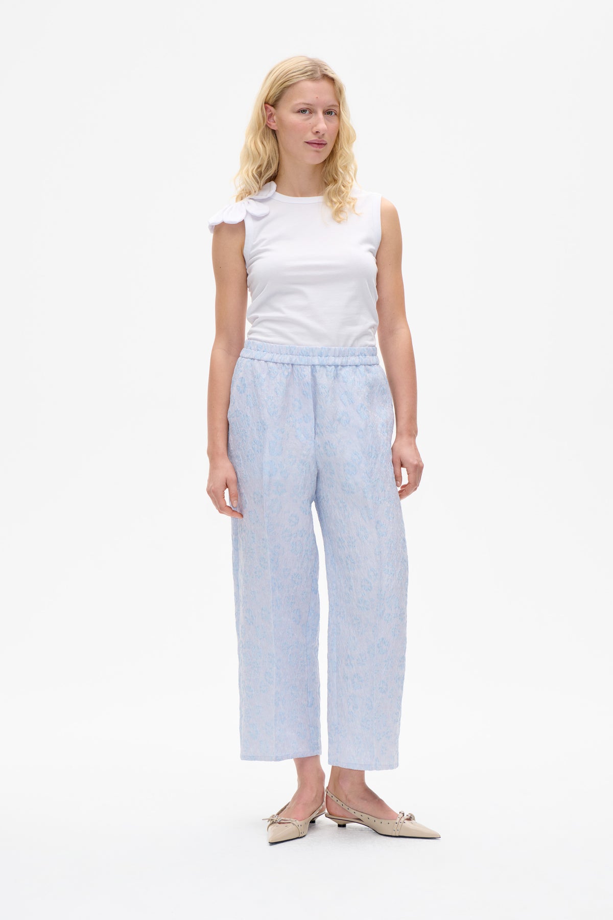 Model wearing a white sleeveless top with a layered flower detail at the shoulder and pale blue patterned wide-leg trousers.
