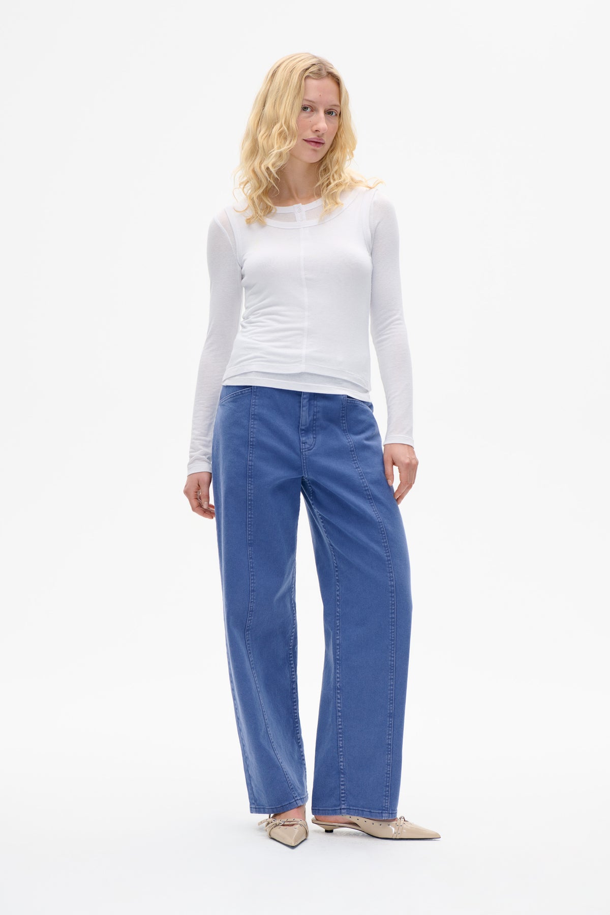 Model wearing a white layered long-sleeve top with a sheer buttoned neckline and blue wide-leg trousers, front view.