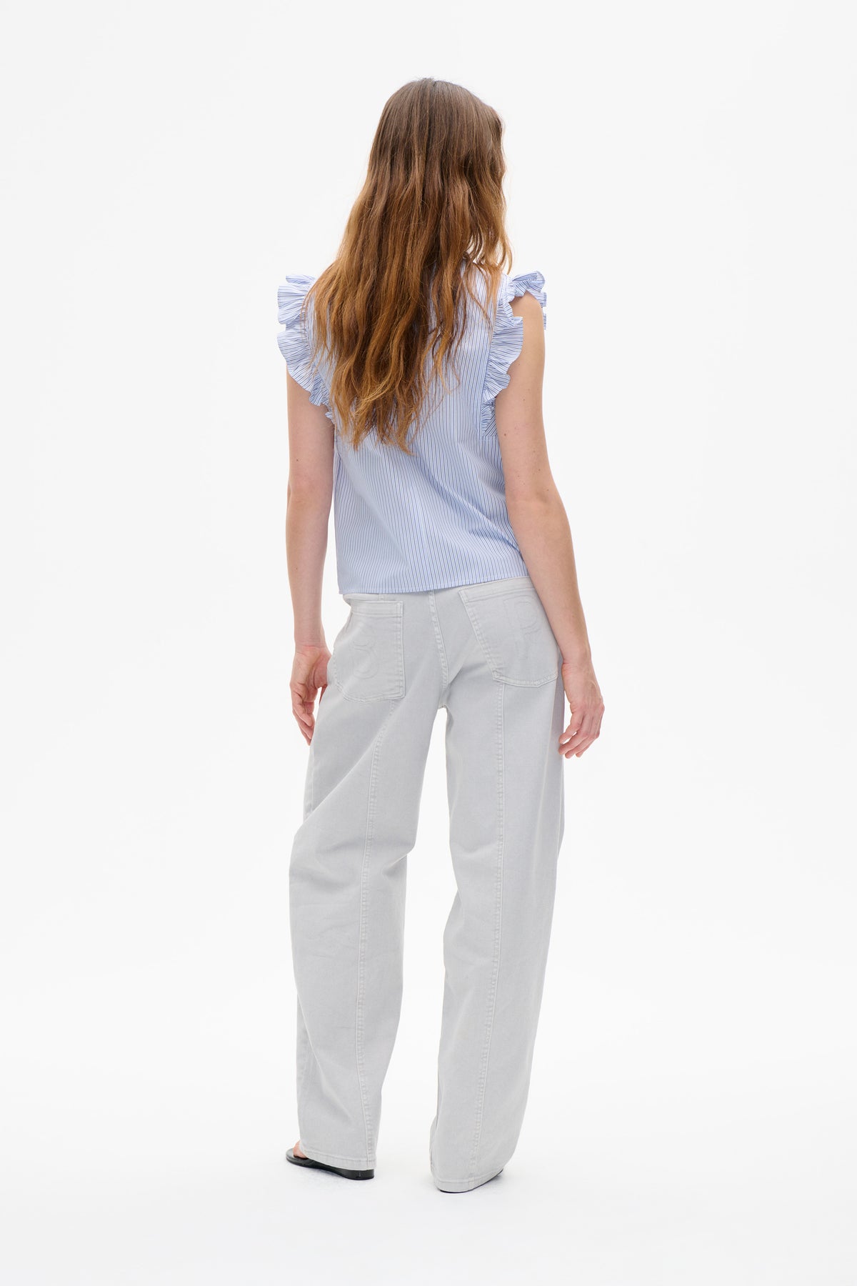Back view of a model wearing a pale blue and white pinstriped sleeveless top with ruffled shoulders and light grey jeans