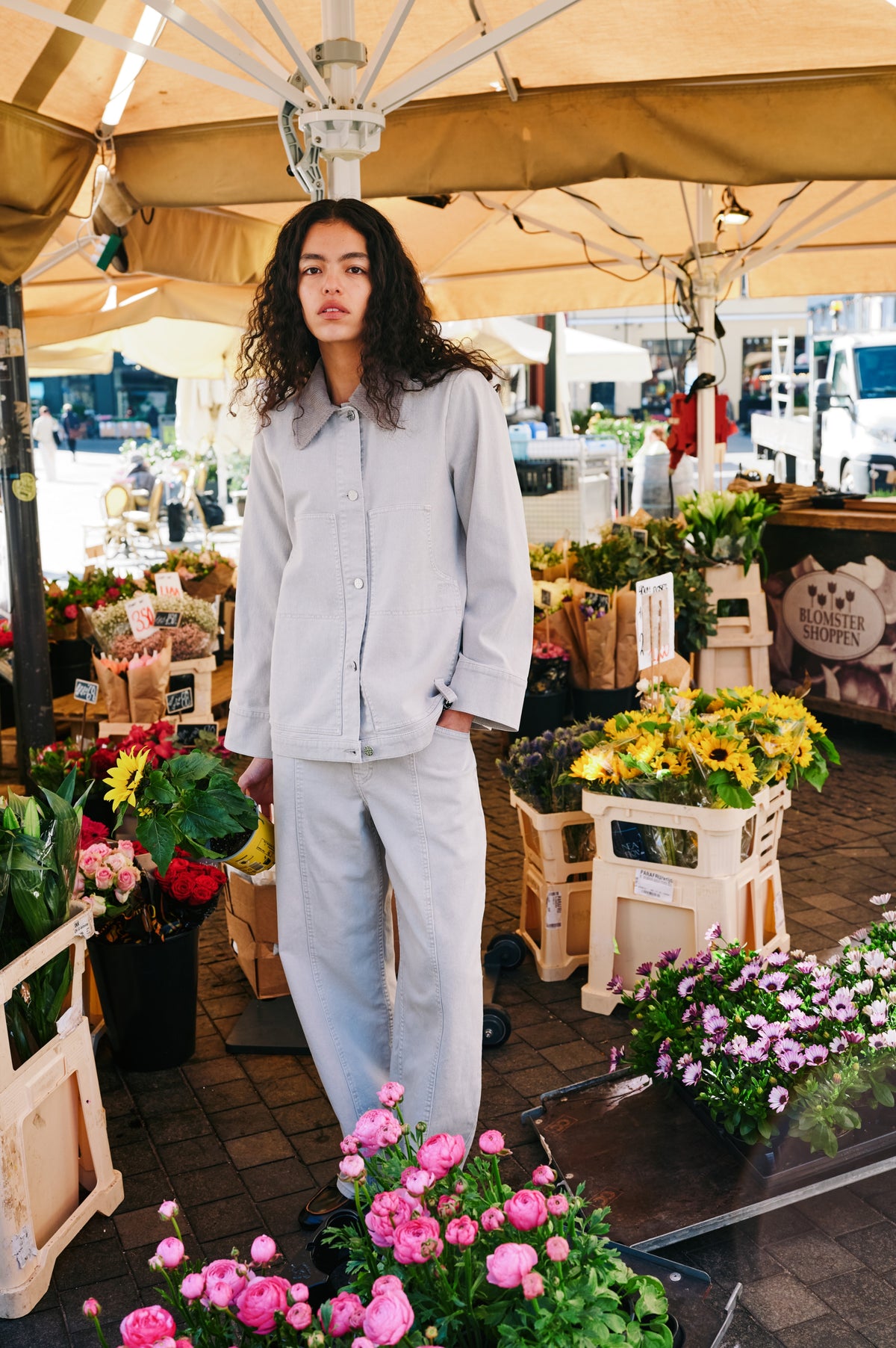 Model wearing a pale blue jacket with a wide ribbed collar and matching jeans among colourful flower stalls.