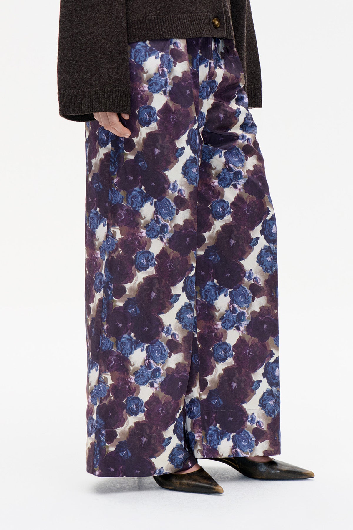 Close-up van een broek met wijde pijpen, met een crème basis en een grote bloemenprint in blauw en pruim, gecombineerd met een donker gebreid vest.