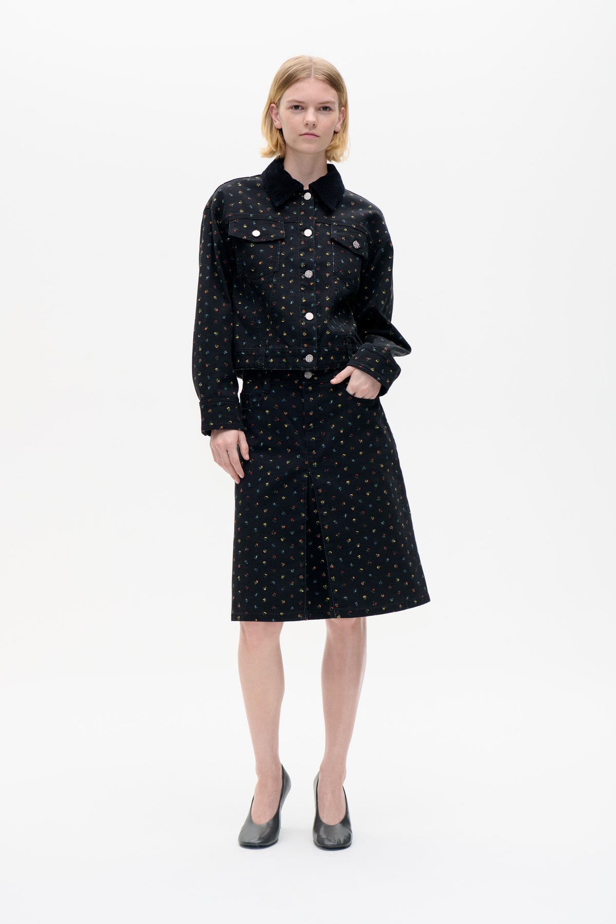 Front view of a model in a black jacket with a dark collar and matching knee-length skirt, both patterned with tiny multicoloured flowers.