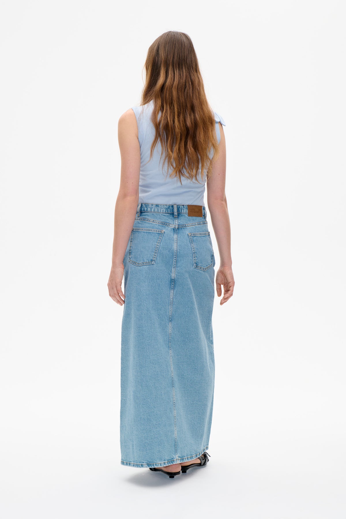 Achteraanzicht van een model in een lichtblauwe mouwloze top met schouderbandjes met strik en een lange lichtgewassen denimrok met opgestikte zakken.