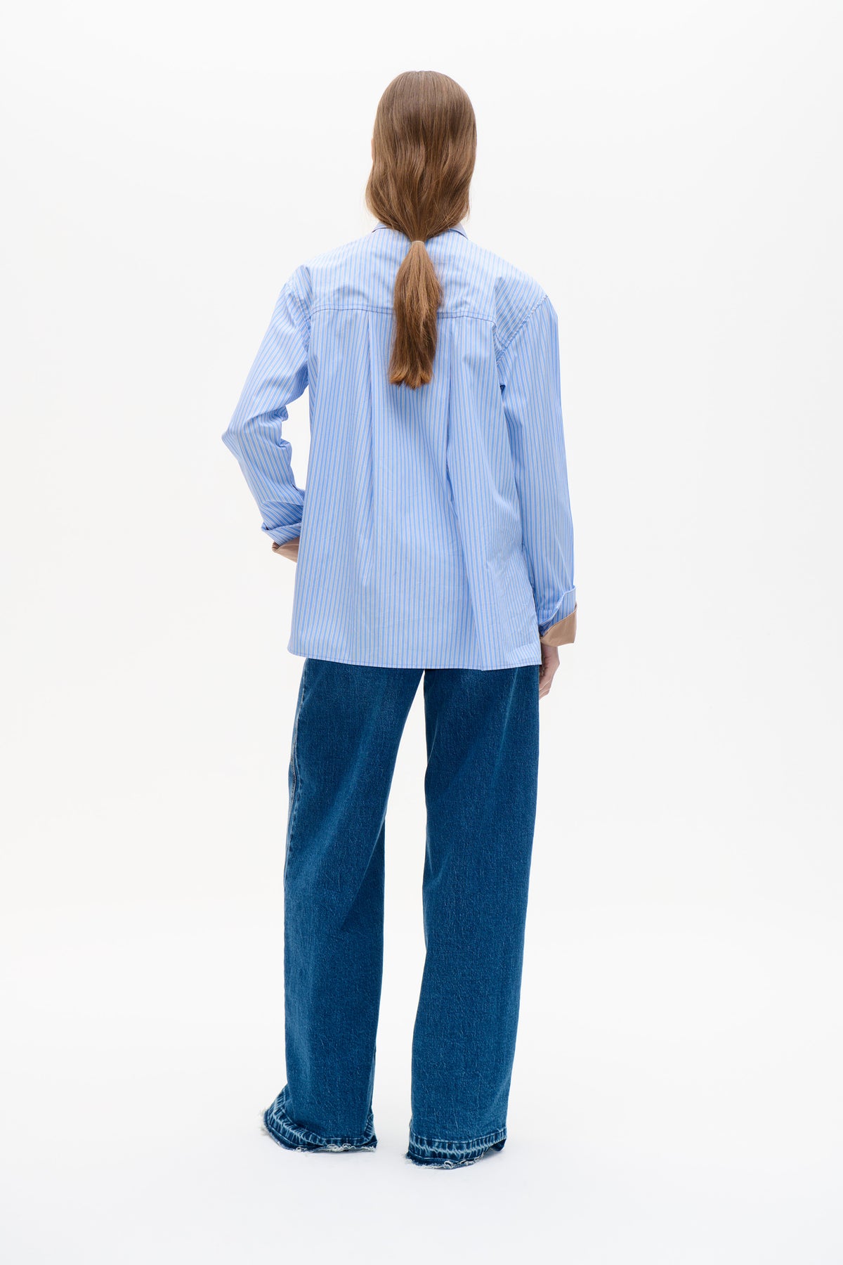 Back view of a model wearing a loose blue-and-white striped long-sleeved shirt with dark wide-leg jeans.