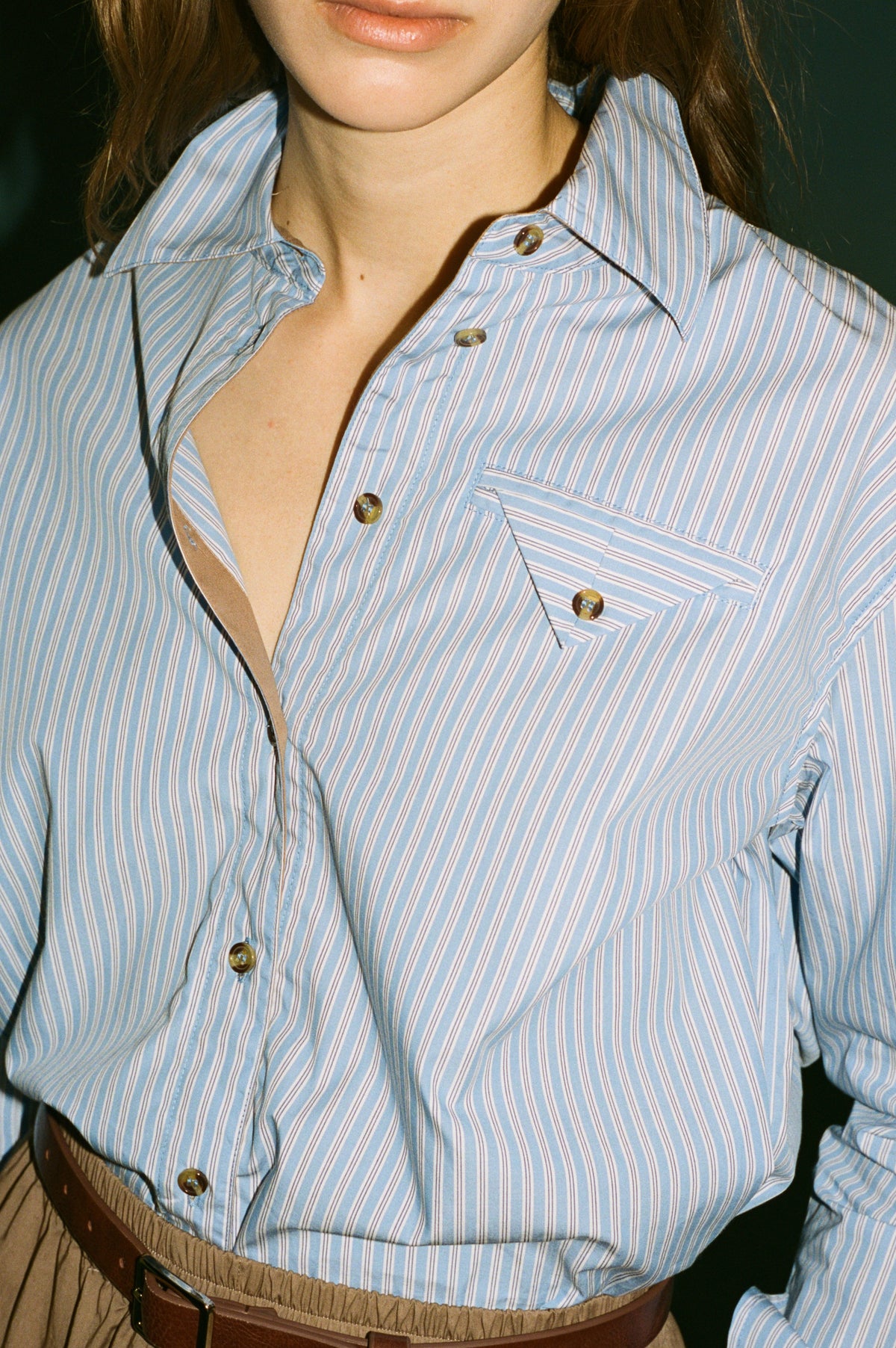 Campaign close-up of a model wearing a blue striped button-down shirt with a chest pocket, tucked into tan trousers with a brown belt.