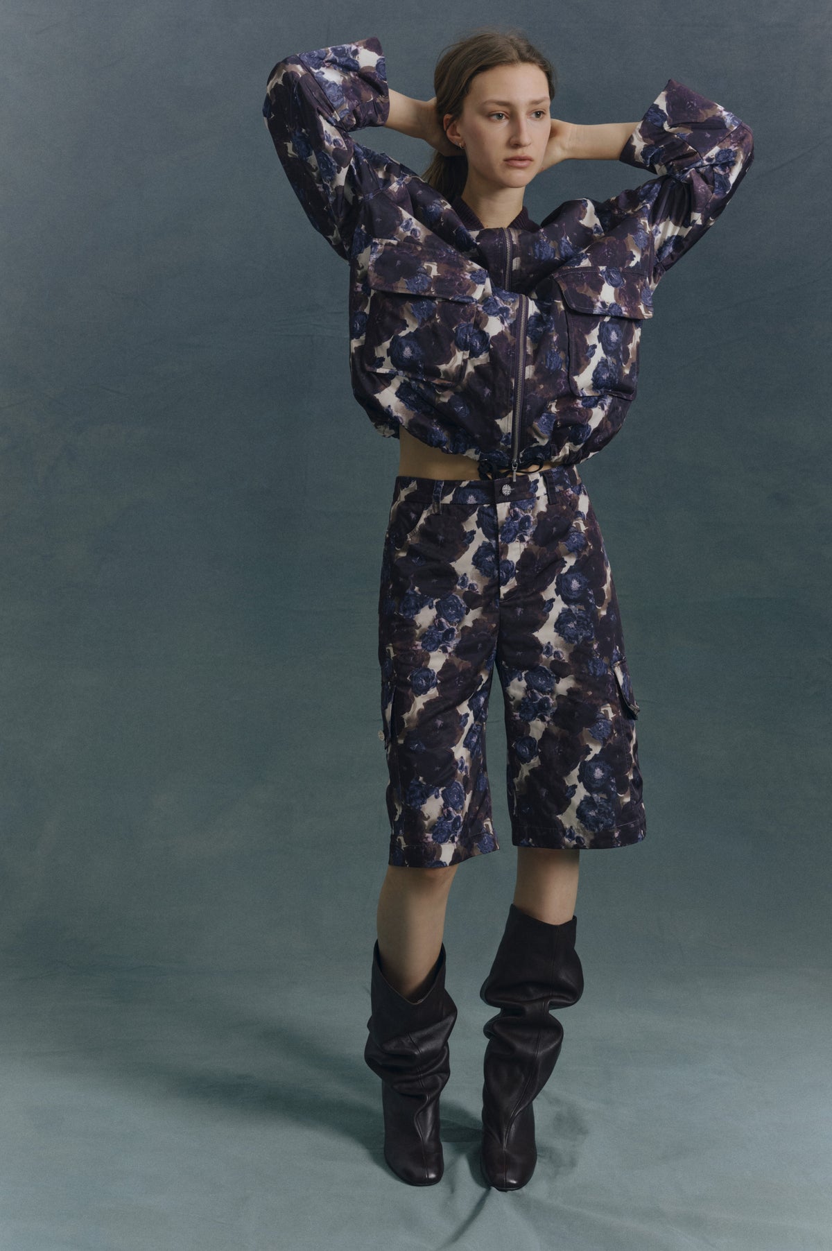 Model poses in a purple and blue floral zip jacket with matching shorts and black knee-high boots against a muted teal backdrop.