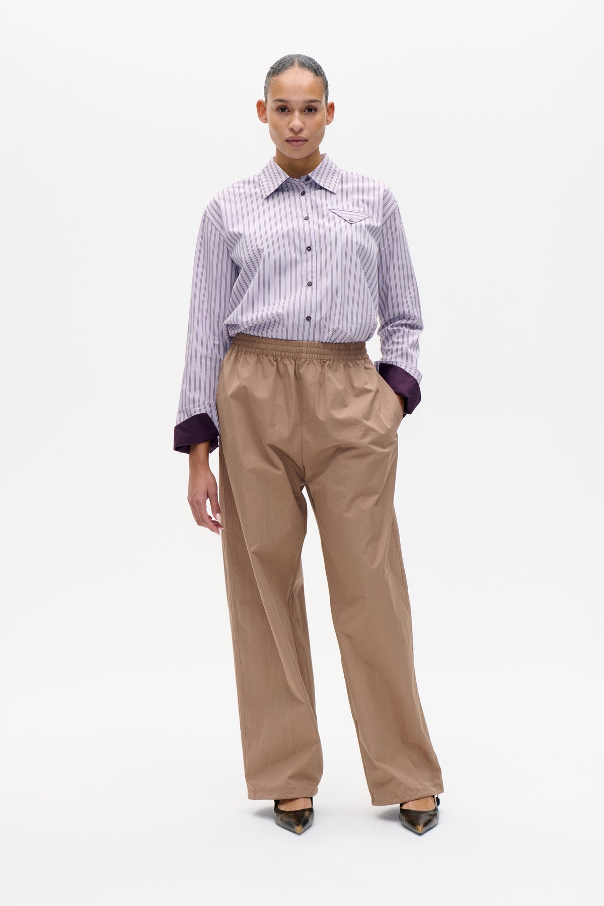 Vooraanzicht van een model dat een lichtlila gestreepte blouse met knoopsluiting, puntkraag en borstzak draagt, gecombineerd met een losse beige broek.
