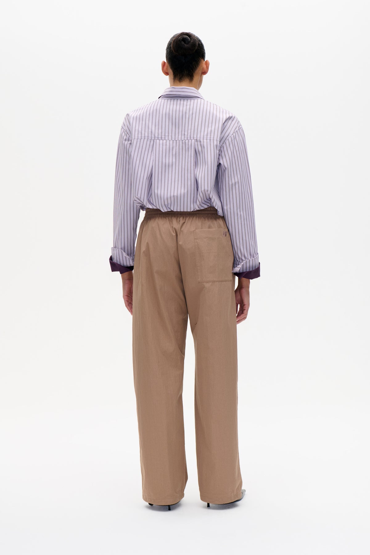 Achteraanzicht van een licht lavendelkleurige blouse met fijne pinstrepen, lange mouwen en contrasterende donkere manchetten, gecombineerd met een zandkleurige broek met brede pijpen.