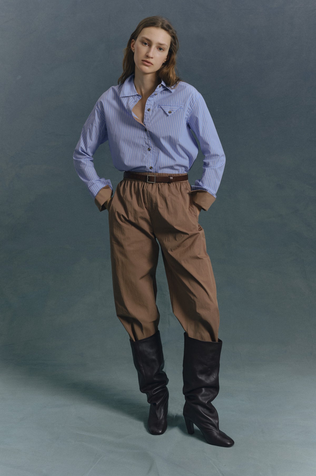 Model draagt een blauw-wit gestreepte blouse met knoopsluiting en borstzak, een bruine broek, een riem en hoge donkere laarzen tegen een petrolkleurige studioachtergrond.
