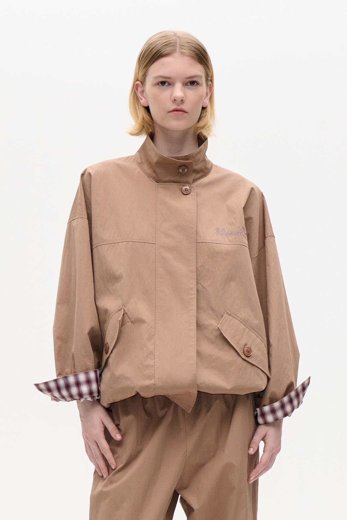 Front view of a tan button-front jacket with a high collar, angled flap pockets, embroidered chest logo and burgundy plaid cuffs.