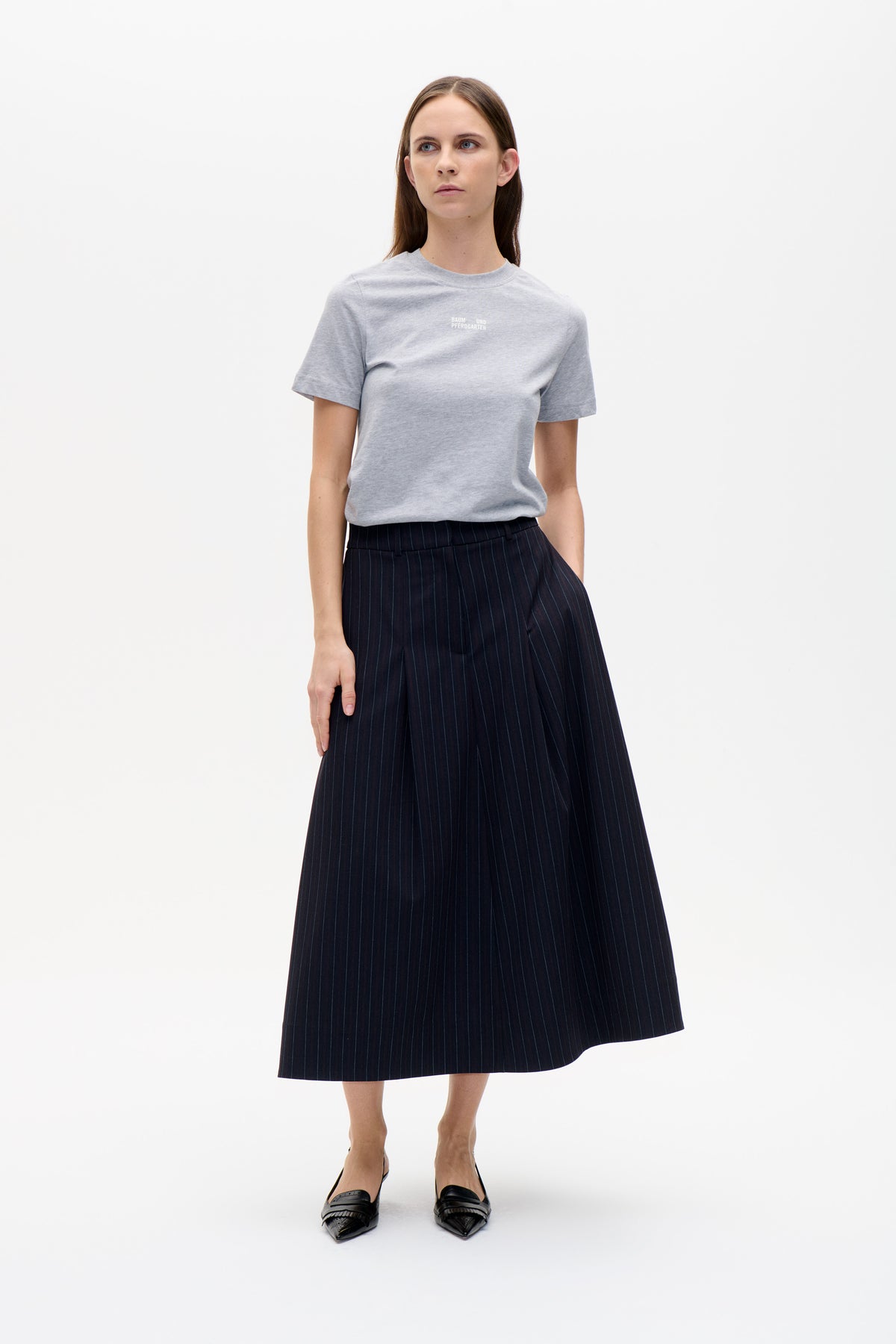 Front view of a light grey short-sleeved T-shirt with a small white chest logo, paired with a dark pinstriped midi skirt.
