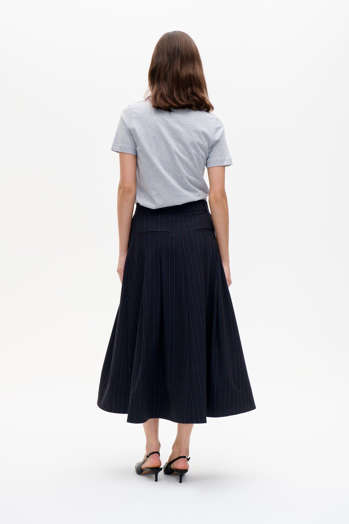 Back view of a model wearing a light grey short-sleeved T-shirt and a dark pinstriped midi skirt with back pockets.