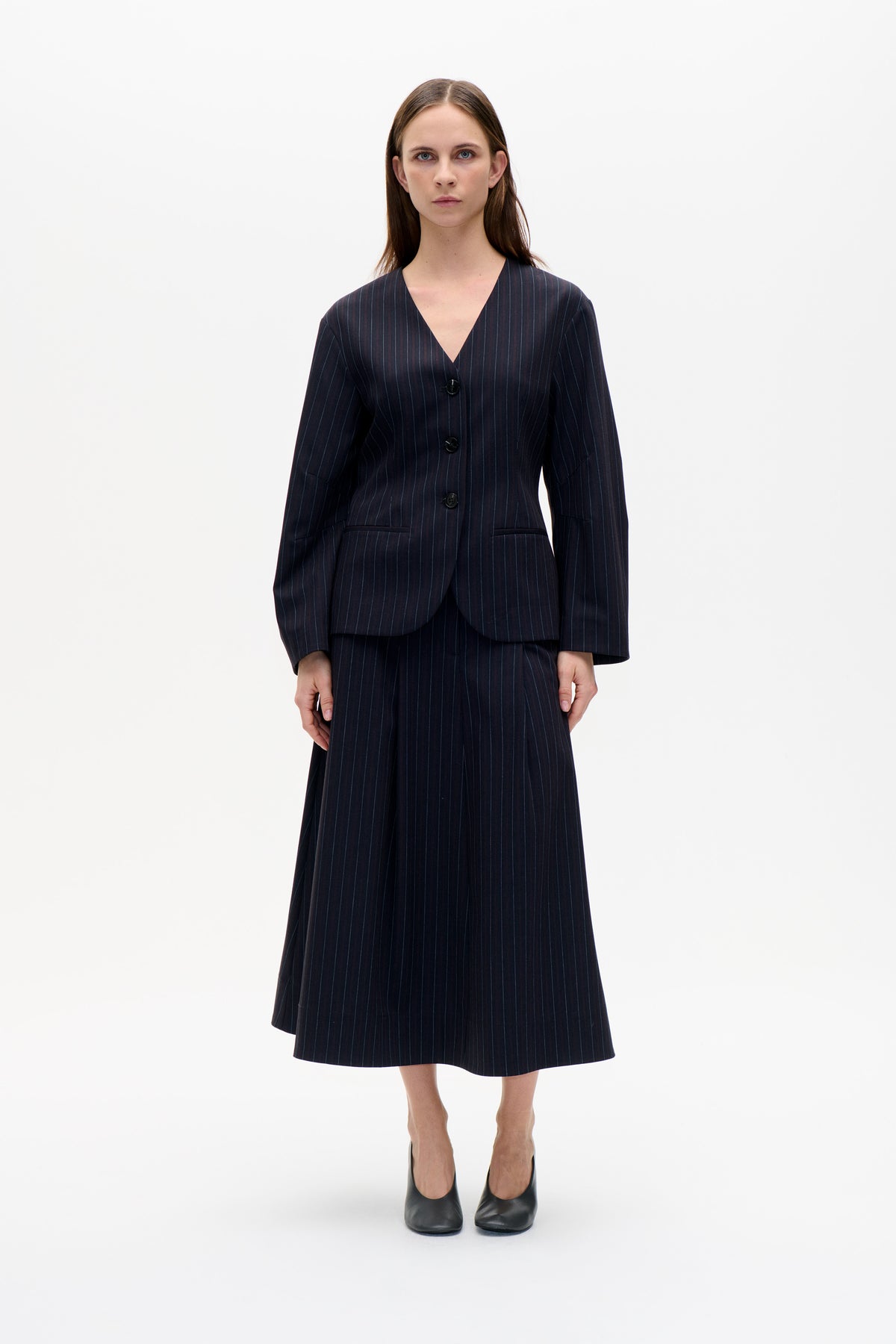 Front view of a model wearing a dark pinstripe V-neck blazer with buttons and welt pockets, paired with a matching knee-length skirt.