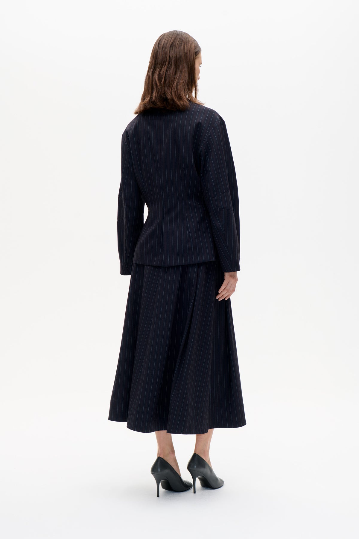 Back view of a model wearing a dark navy pinstripe blazer and matching midi skirt with black heels.