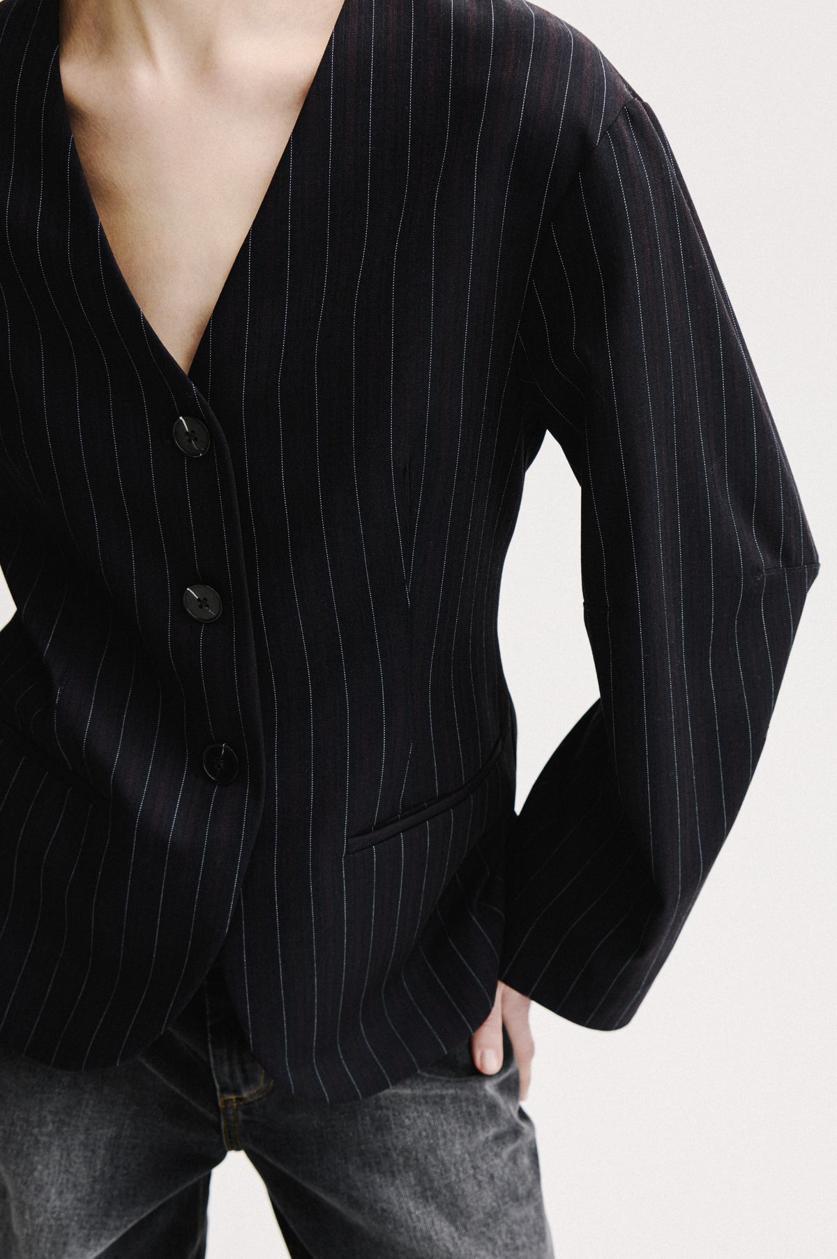 Close-up campaign view of a black pinstripe blazer with a deep V-neck, dark buttons and a welt pocket, paired with grey jeans.