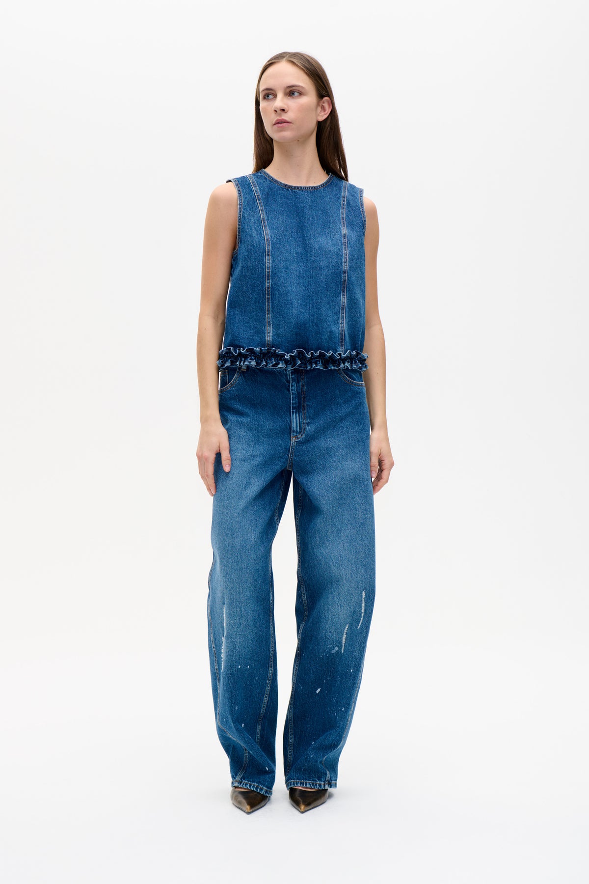 Model wearing a blue sleeveless denim top with vertical seams and a ruffled hem, paired with blue jeans featuring white distressed marks.