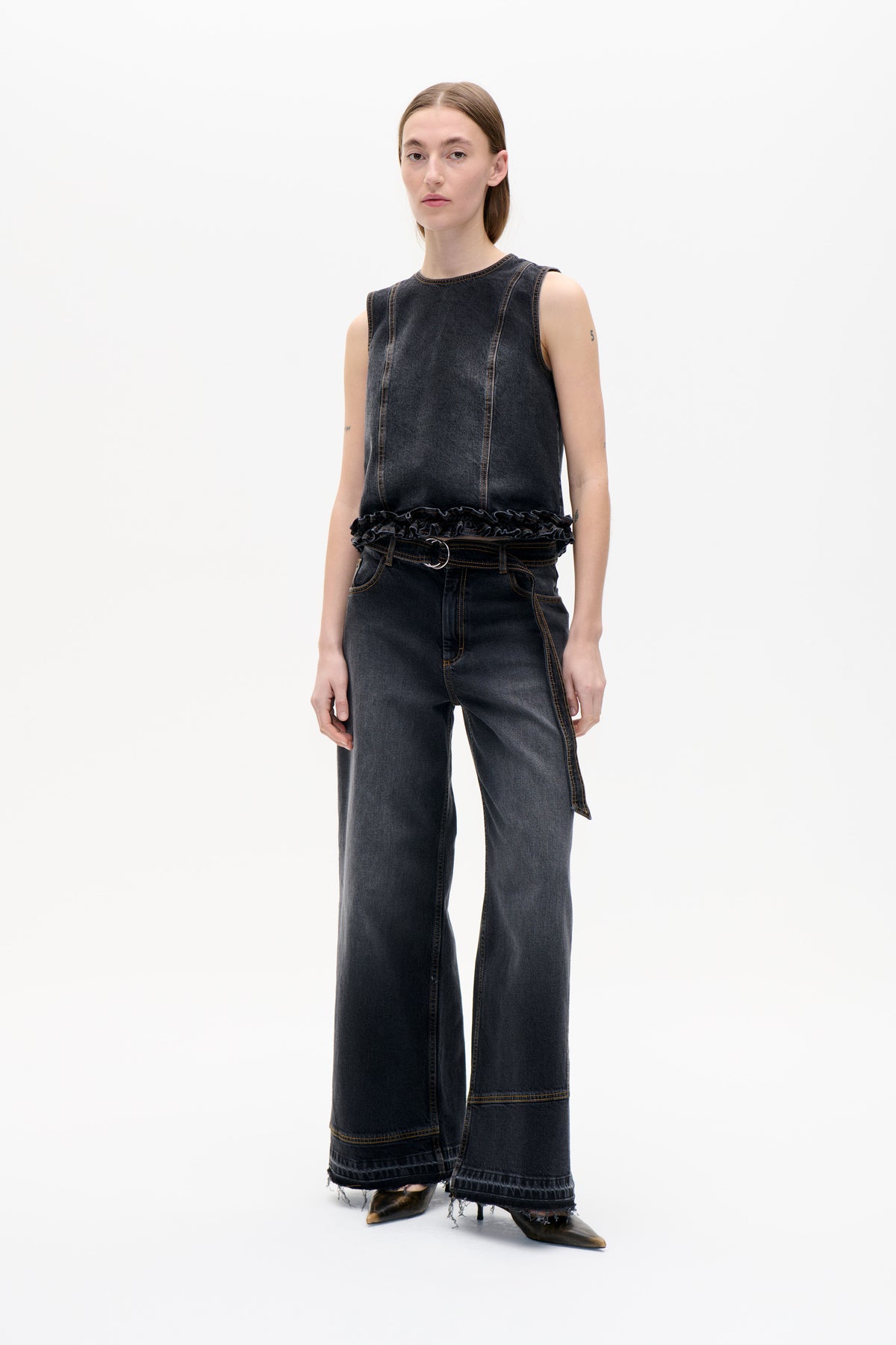 Front view of a model wearing a charcoal denim sleeveless top with contrast stitching and a ruffled hem, paired with wide-leg jeans.