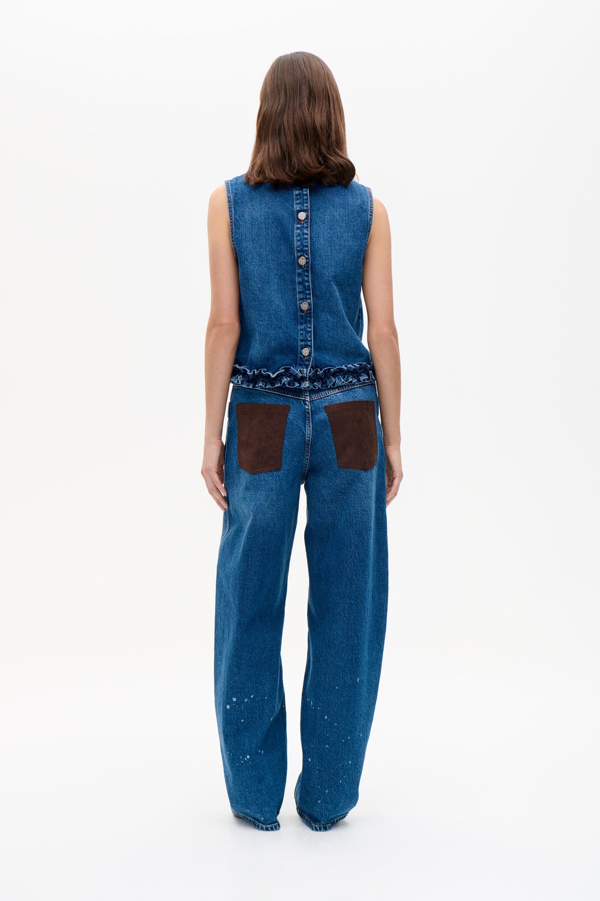 Achteraanzicht van een model dat een blauw denim mouwloos topje draagt met knopen achterop en een gerimpelde zoom, gecombineerd met wijde jeans met bruine opgestikte zakken.