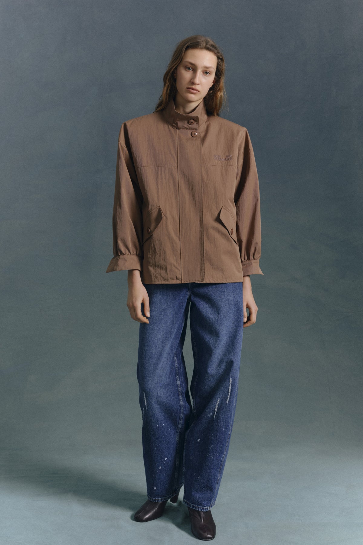 Model draagt een taupe jasje met knoopsluiting, staande kraag en klepzakken, gecombineerd met versleten blauwe jeans tegen een gedempte teal-achtergrond.