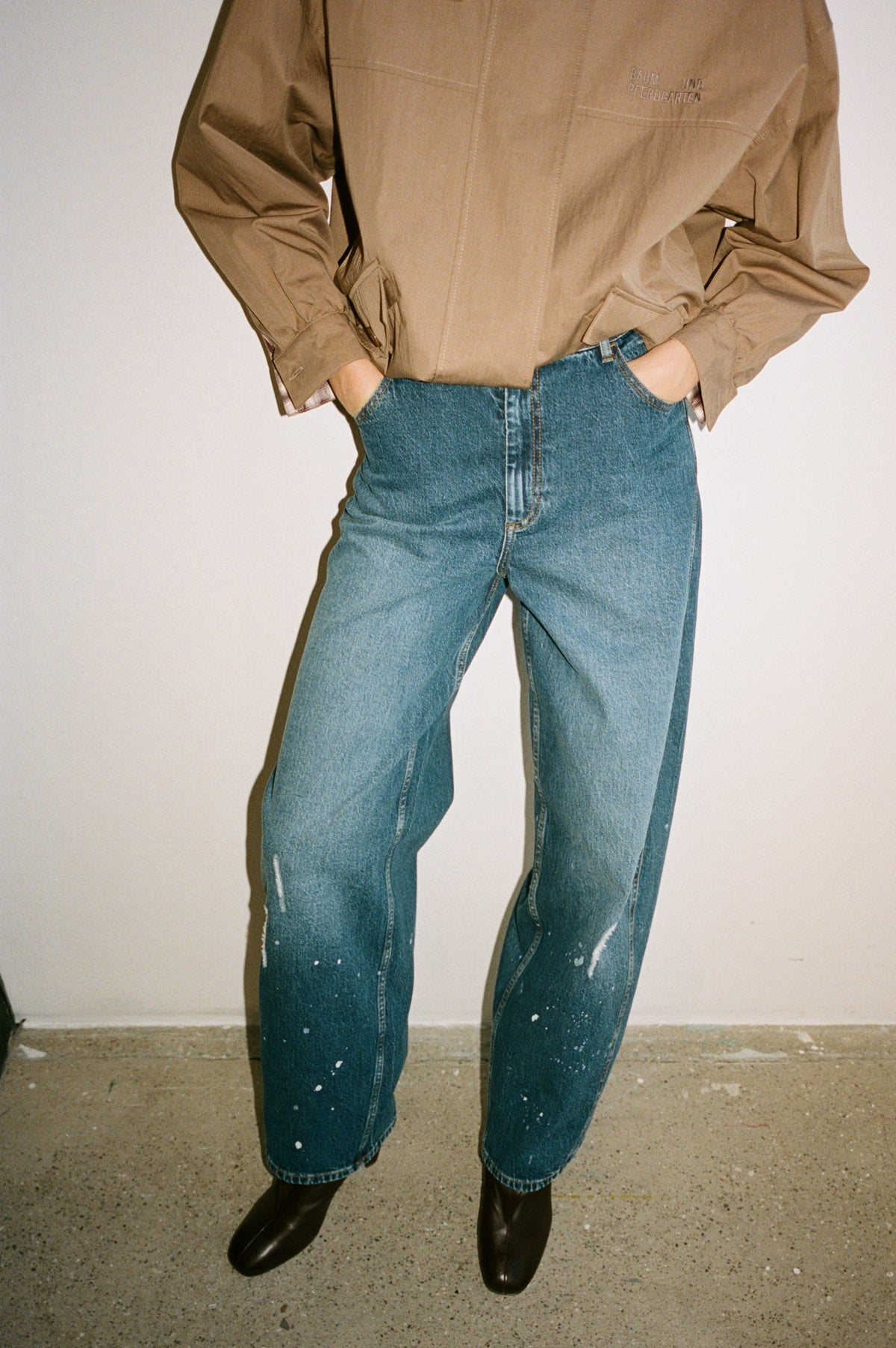 Bijgesneden campagnebeeld van een model in een taupekleurige blouse met een geborduurd logo, blauwe jeans met verfspatten en zwarte laarzen.