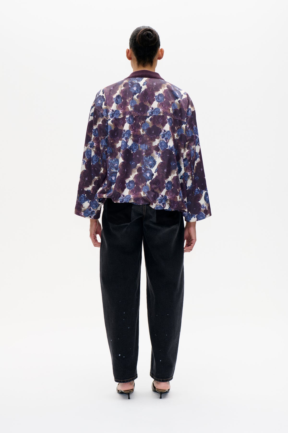 Achteraanzicht van een model dat een crèmekleurige jas draagt met een grote blauw-paarse bloemenprint en een geribde pruimkleurige kraag.