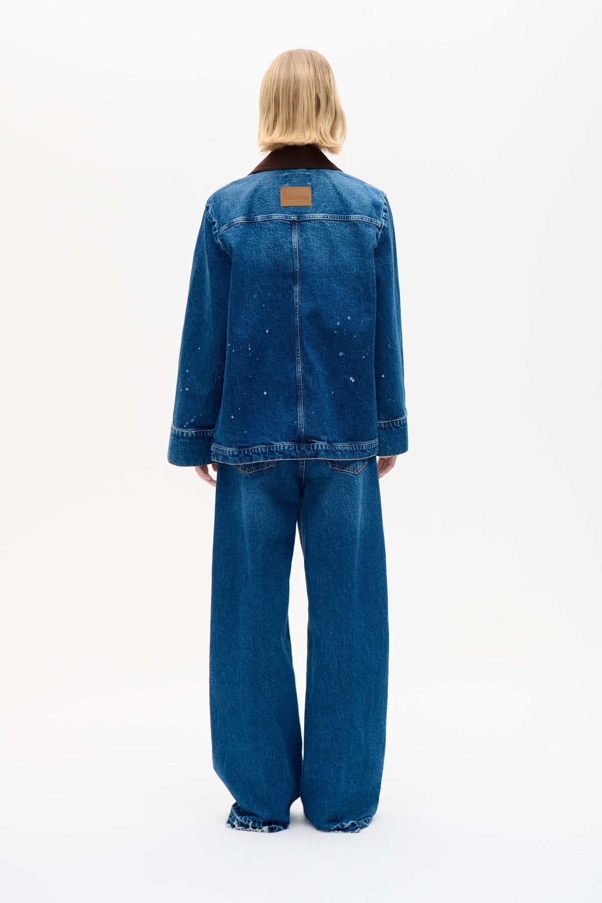 Back view of a model wearing a blue denim jacket with a dark brown collar and scattered pale flecks, paired with wide-leg jeans.