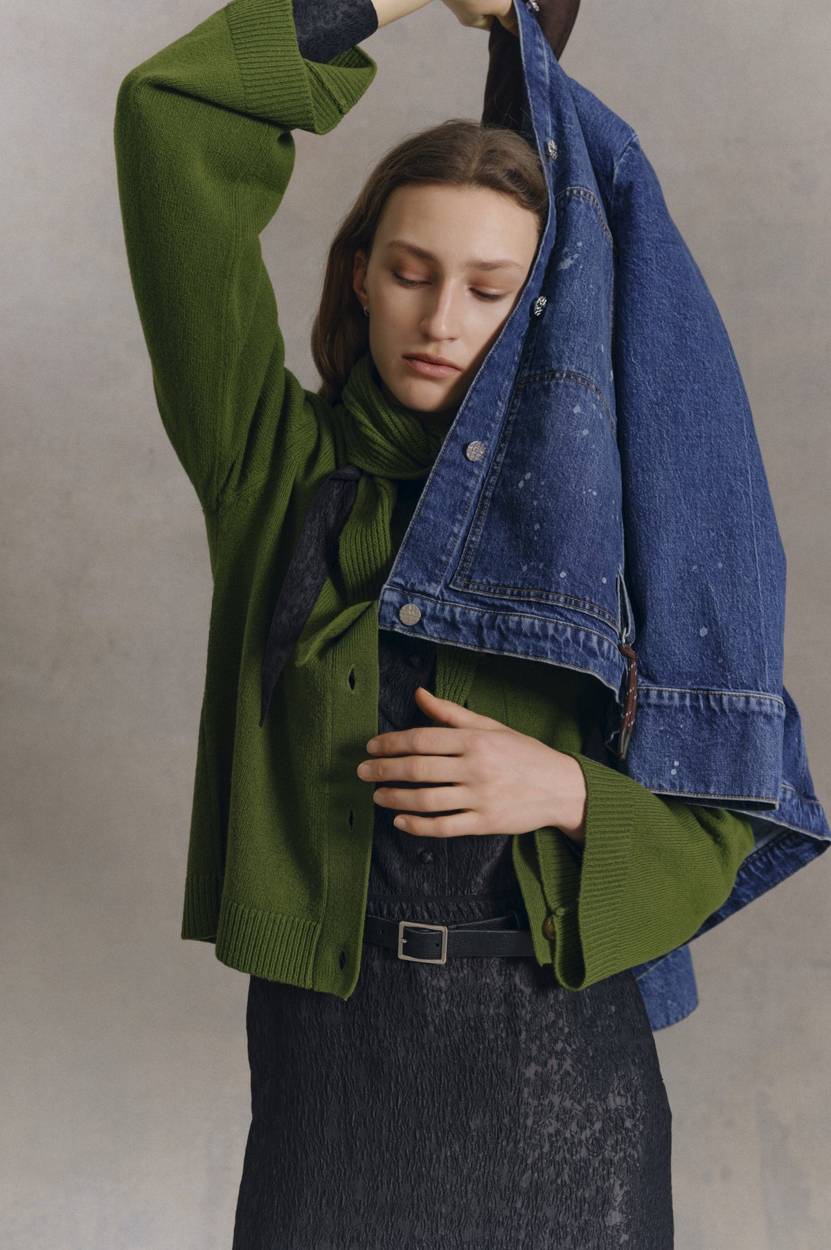 Close-cropped campaign portrait of a model in a green ribbed cardigan and black textured belted dress, holding a mottled blue denim jacket.