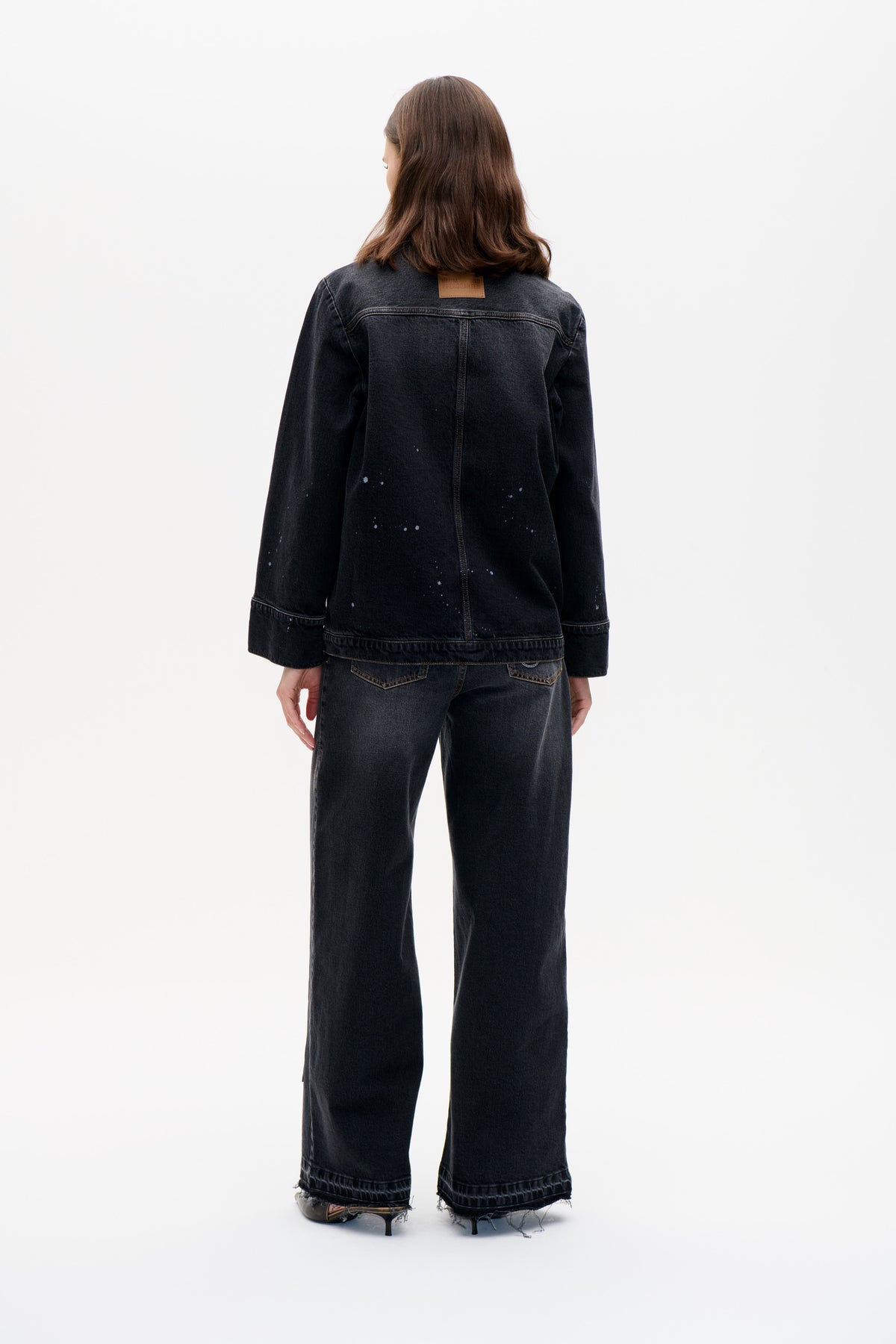 Back view of a model wearing a black denim jacket with scattered pale speckles and matching wide-leg jeans with frayed hems.