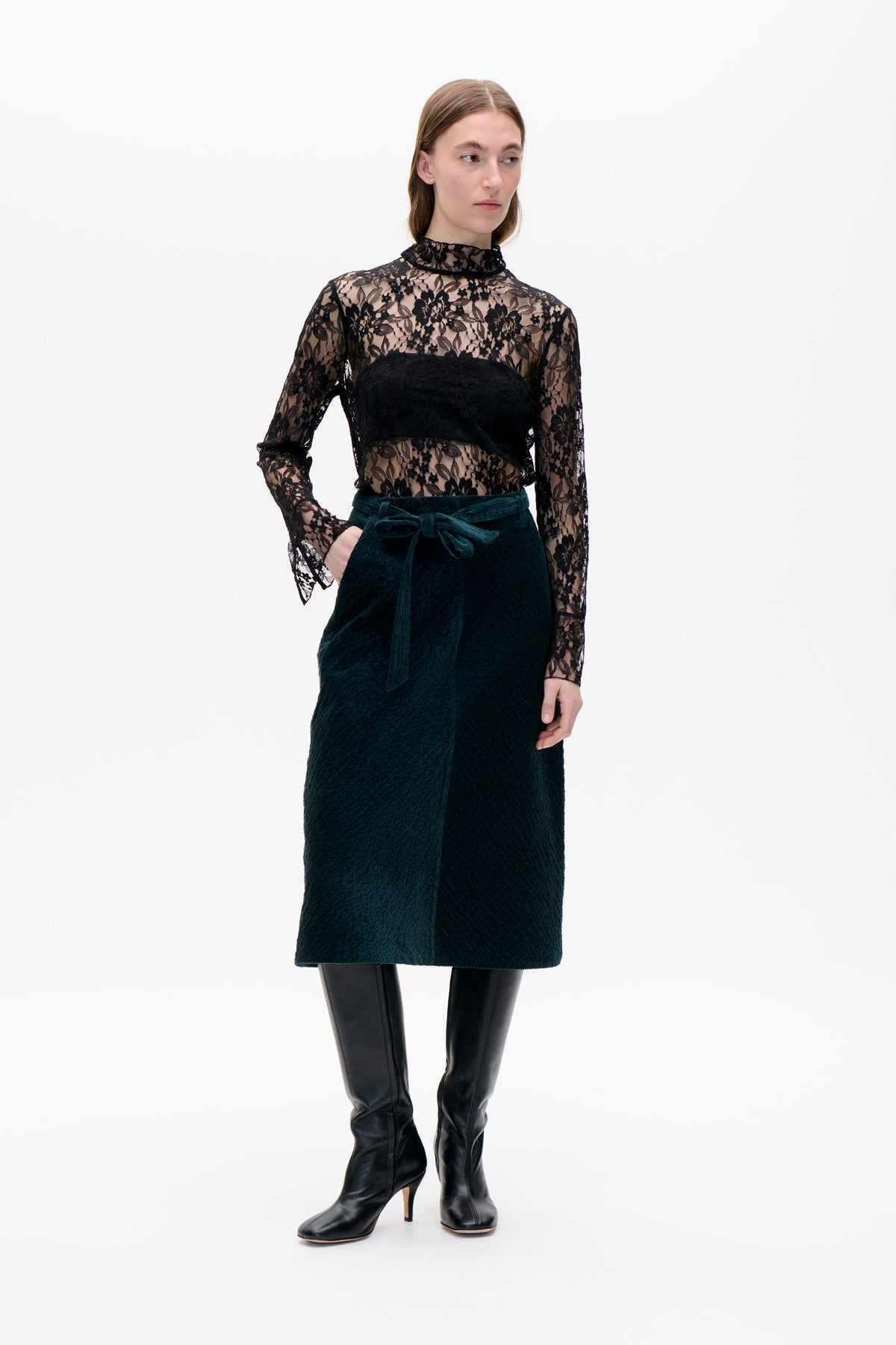 Model trägt ein transparentes schwarzes Spitzenoberteil mit floralem Muster und hohem Kragen sowie blickdichter Brustpartie, einen dunkelpetrolfarbenen, gerippten Rock mit Taillengürtel und schwarze kniehohe Stiefel.