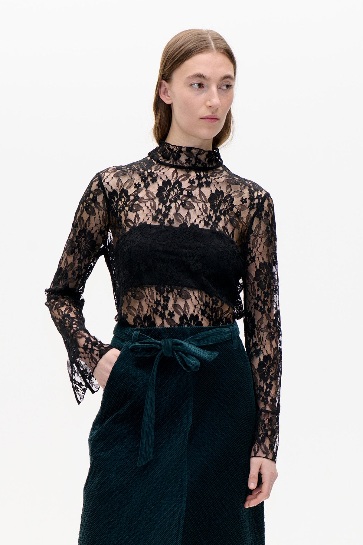 Model trägt ein transparentes schwarzes Spitzenoberteil mit Blumenmuster, hohem Kragen und ausgestellten Manschetten über einem schwarzen Bandeau, kombiniert mit einem dunkelpetrolfarbenen Rock.