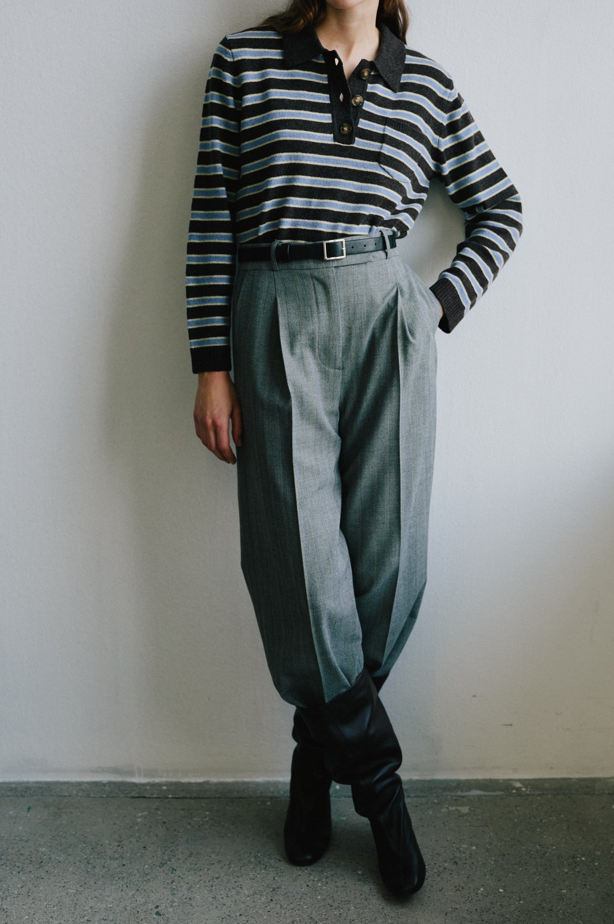 Campaign view of a model in a black knit polo with blue and cream stripes, grey wide-leg trousers, a black belt and boots.