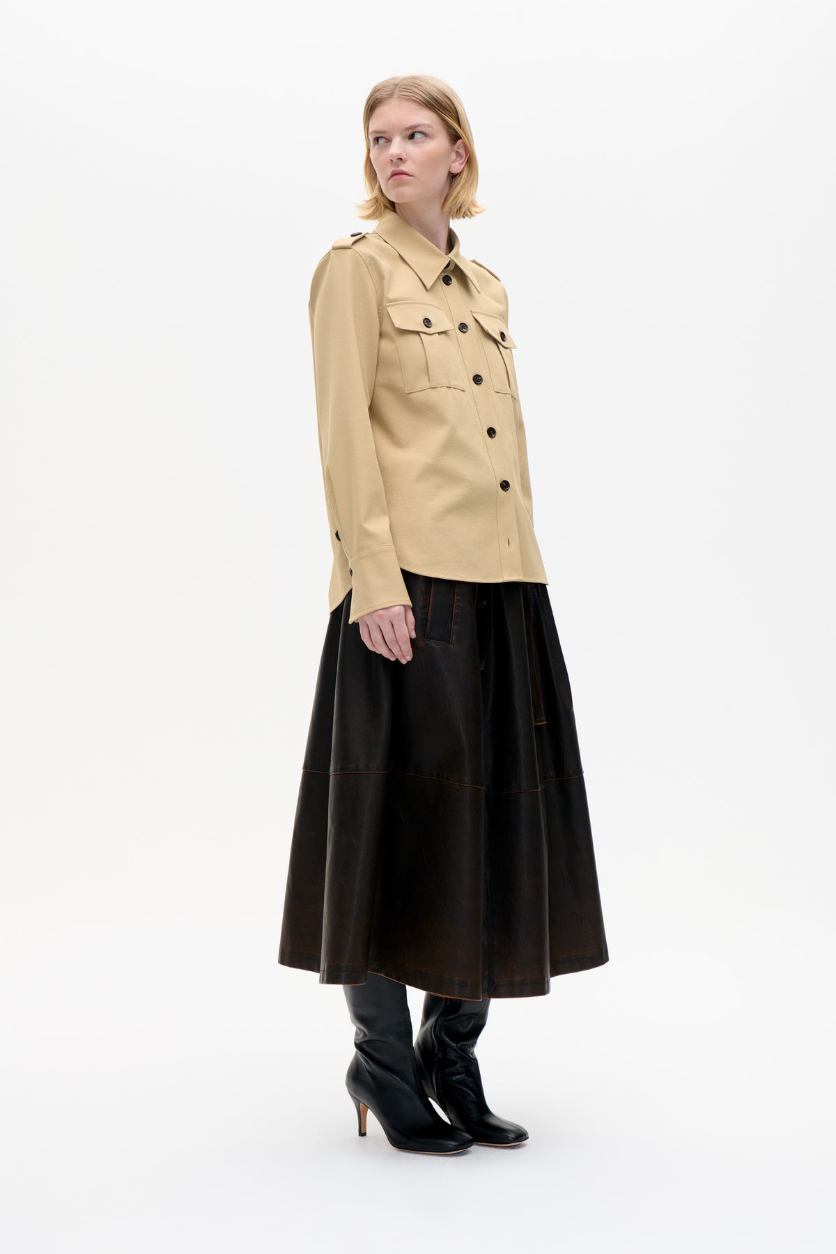 Model wearing a beige button-front shirt jacket with flap chest pockets and shoulder tabs, paired with a black paneled skirt and tall boots.