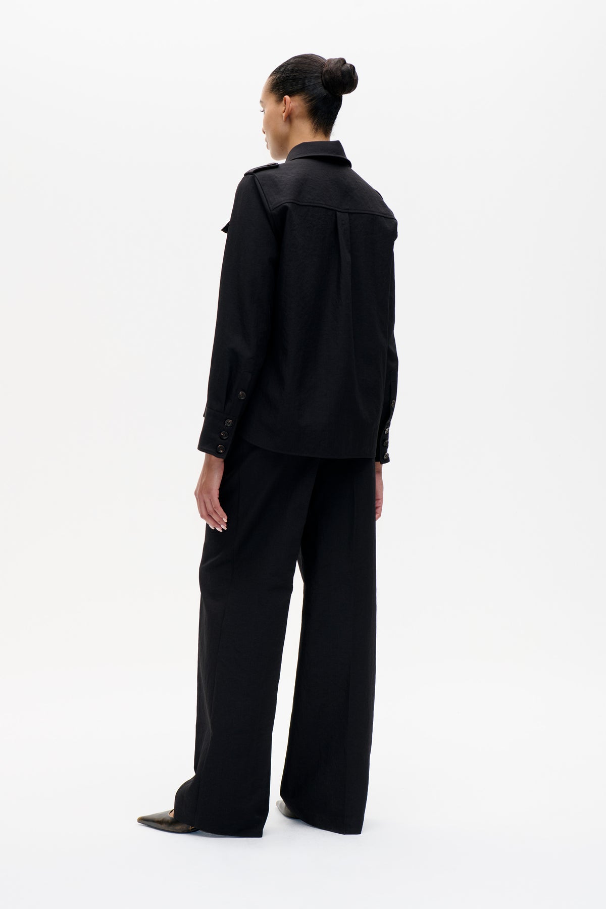 Mannequin vu de dos portant une veste noire à col, avec empiècement au dos et poignets boutonnés, associée à un pantalon à jambes larges.