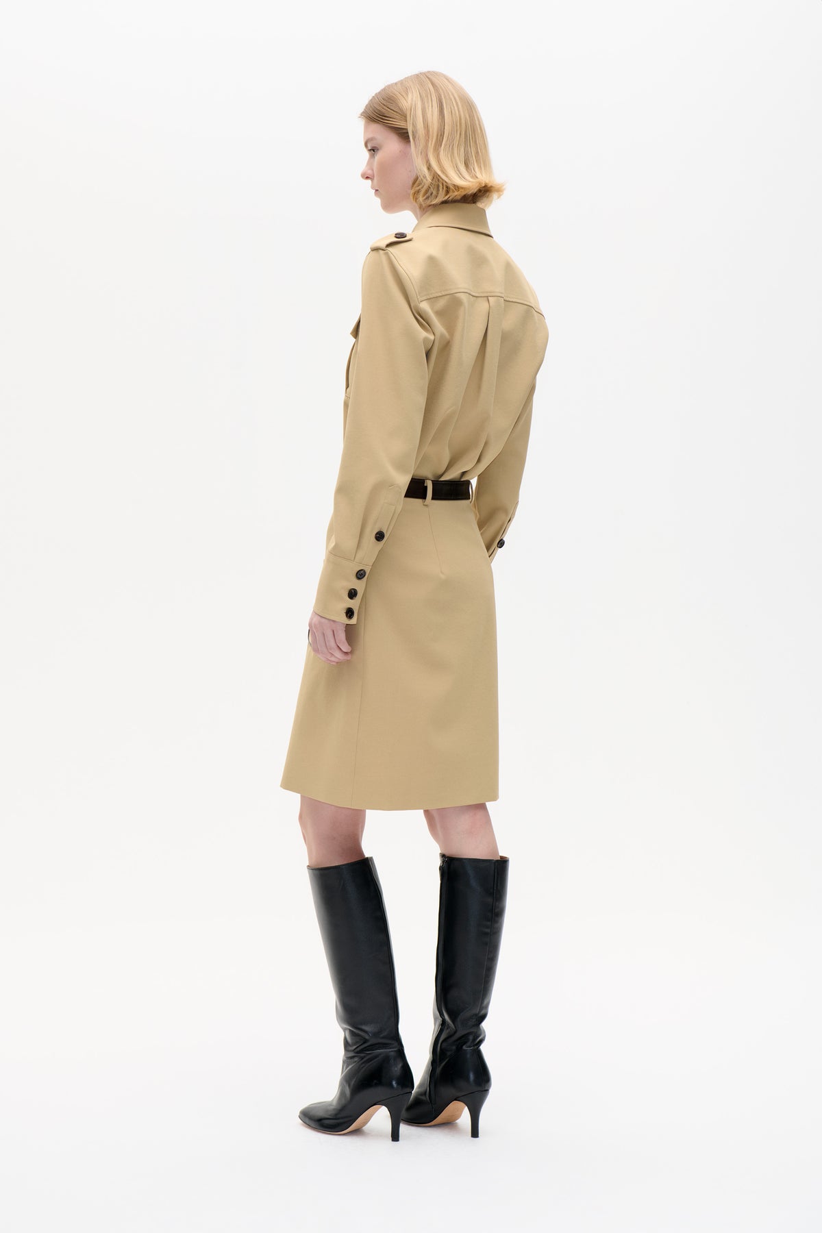 Side-back view of a model wearing a beige collared shirt with a back yoke, tucked into a matching knee-length skirt with a black belt and tall black heeled boots.