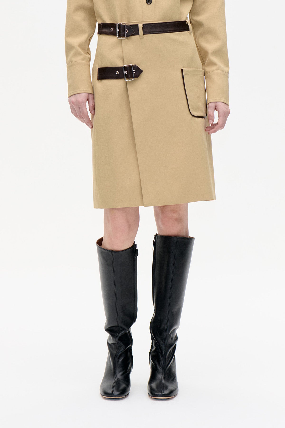 Front view of a beige knee-length coat with two dark buckle straps, a black-edged patch pocket and black knee-high boots.