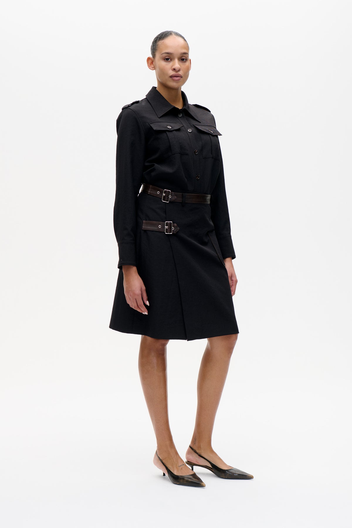 Model wearing a black button-front shirt dress with flap pockets, shoulder tabs and two dark brown buckled belts.