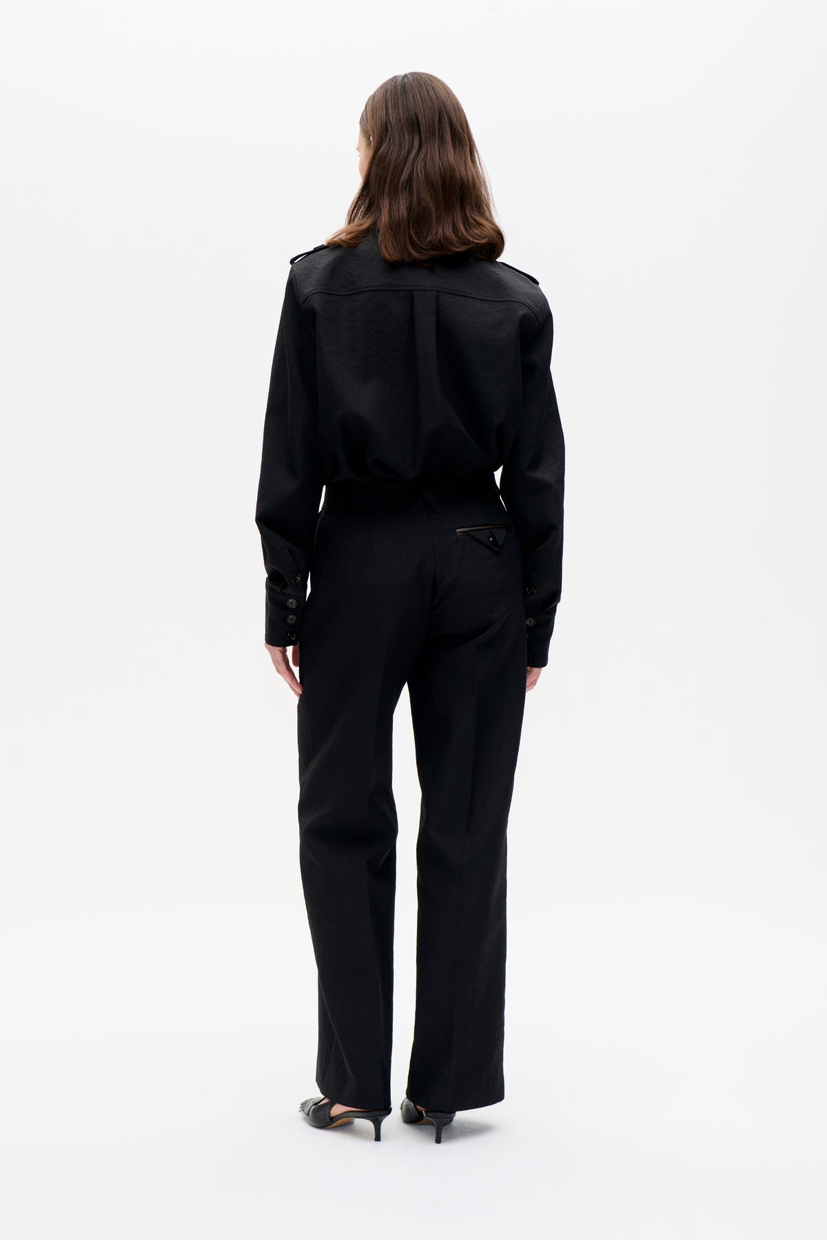 Back view of a model wearing a black belted jacket with shoulder tabs and button cuffs, paired with wide-leg trousers.