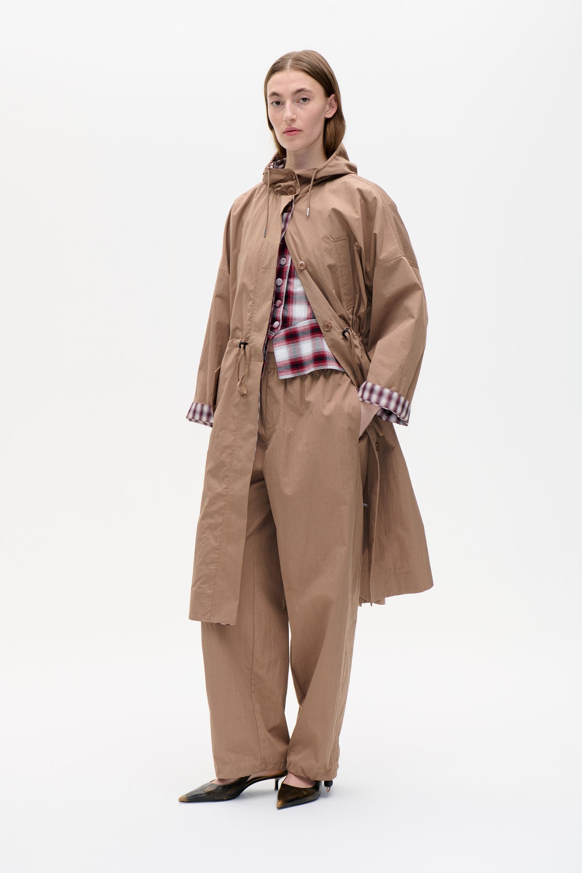 Model wearing a taupe hooded long coat with button fastening over a red, white and navy plaid shirt and matching trousers.