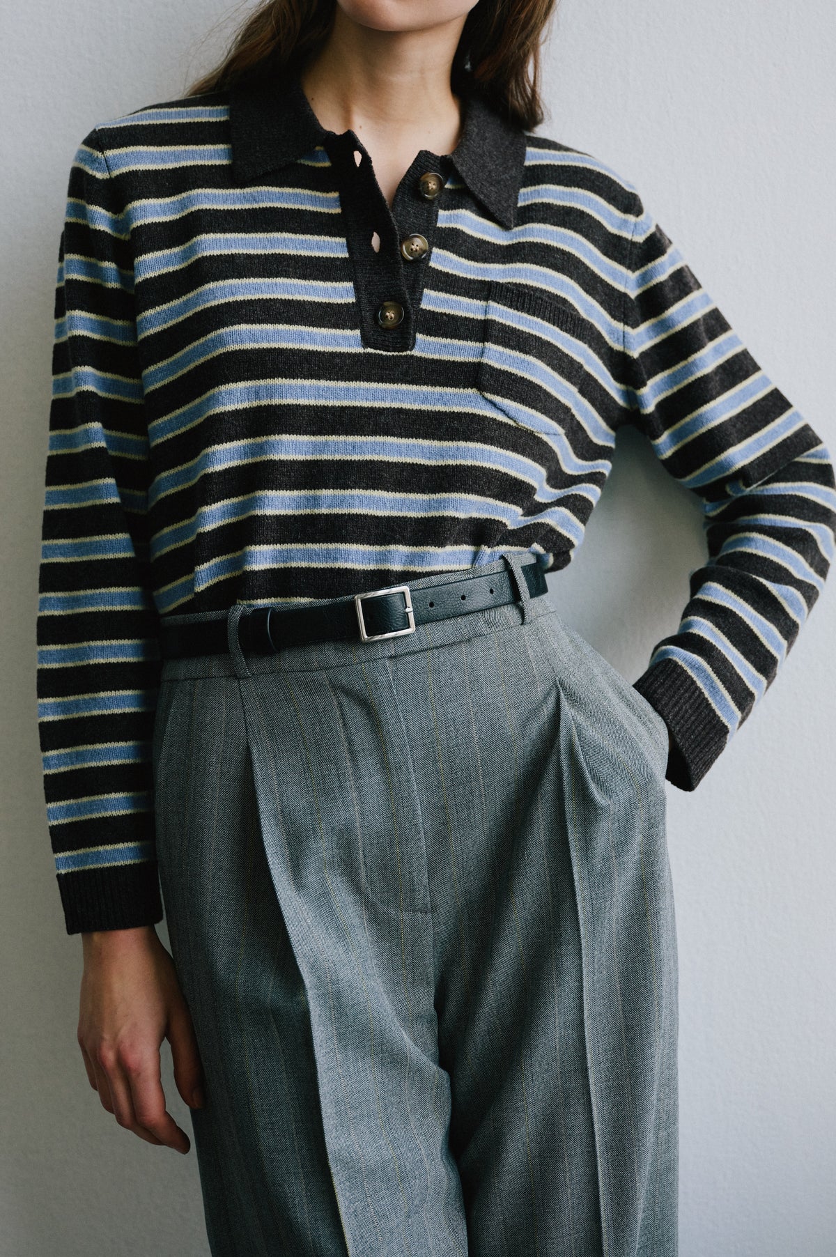 Close-up of a charcoal knit long-sleeve polo with blue and cream stripes, styled with grey checked trousers and a black belt.