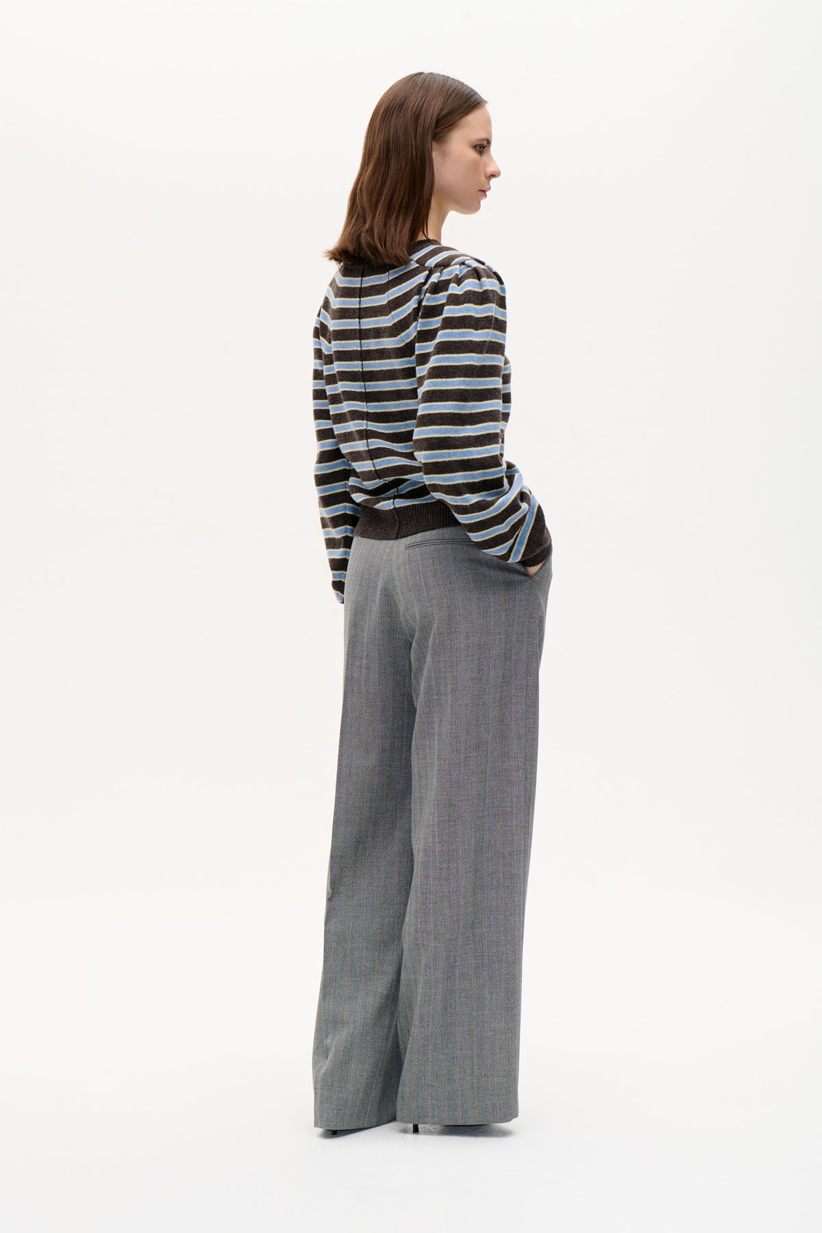 Side-profile model wearing a dark brown sweater with blue and cream stripes and grey wide-leg trousers against a plain backdrop.