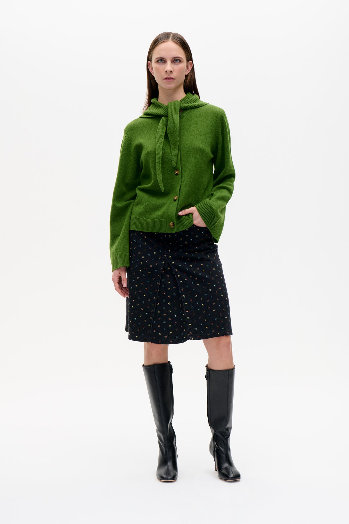 Model wearing a bright green button-front cardigan with a matching knit scarf, a black skirt with tiny multicoloured flowers, and tall black boots.