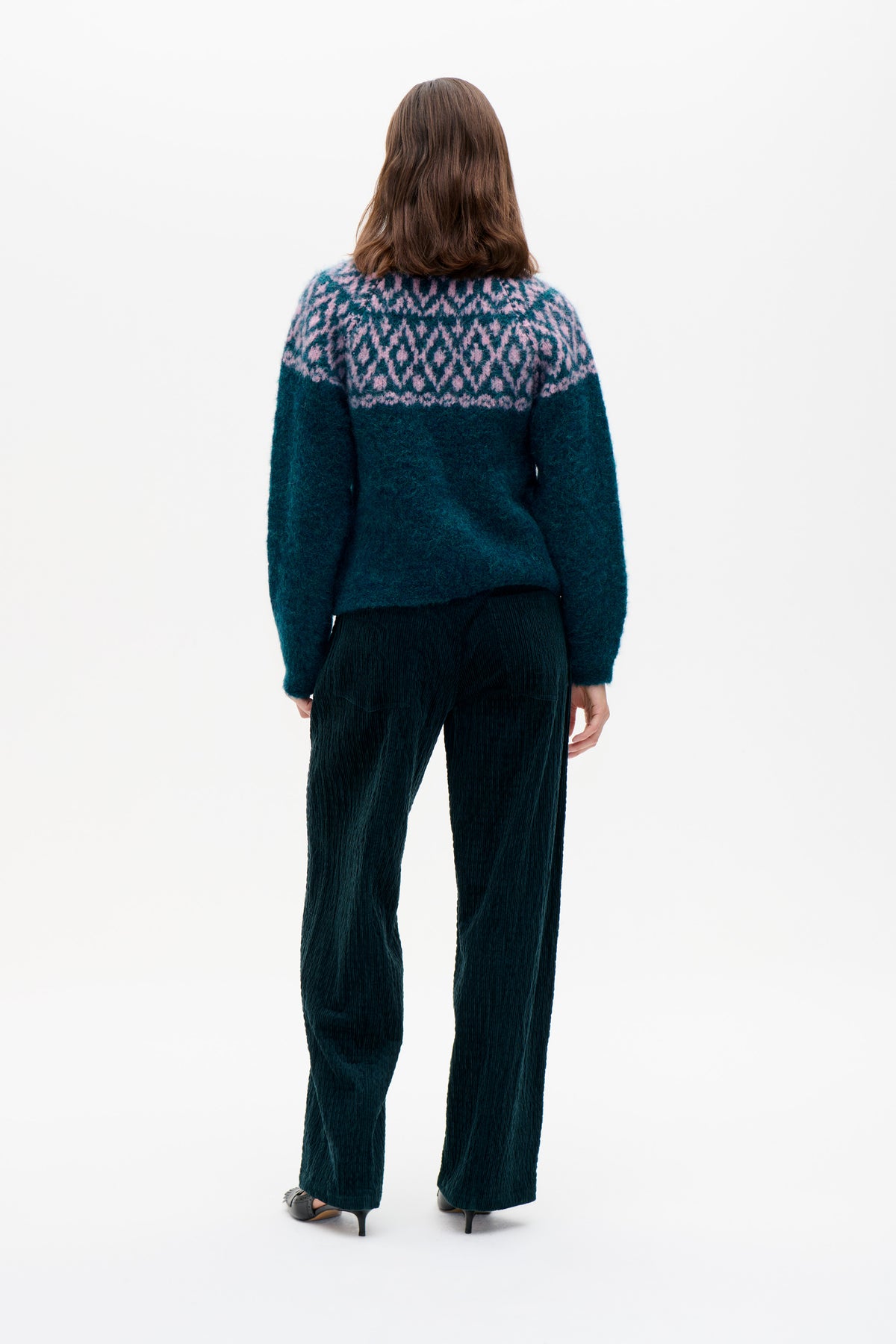 Back view of a model wearing a dark teal knit sweater with a pink geometric yoke pattern and dark corduroy trousers.
