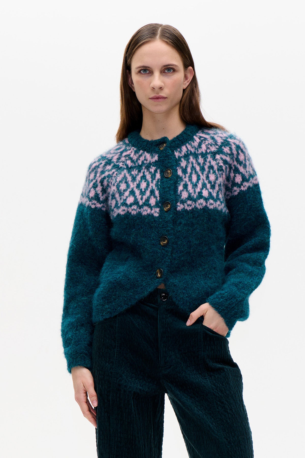 Model wearing a teal button-front cardigan with a pink and pale blue geometric yoke pattern, paired with dark corduroy trousers.
