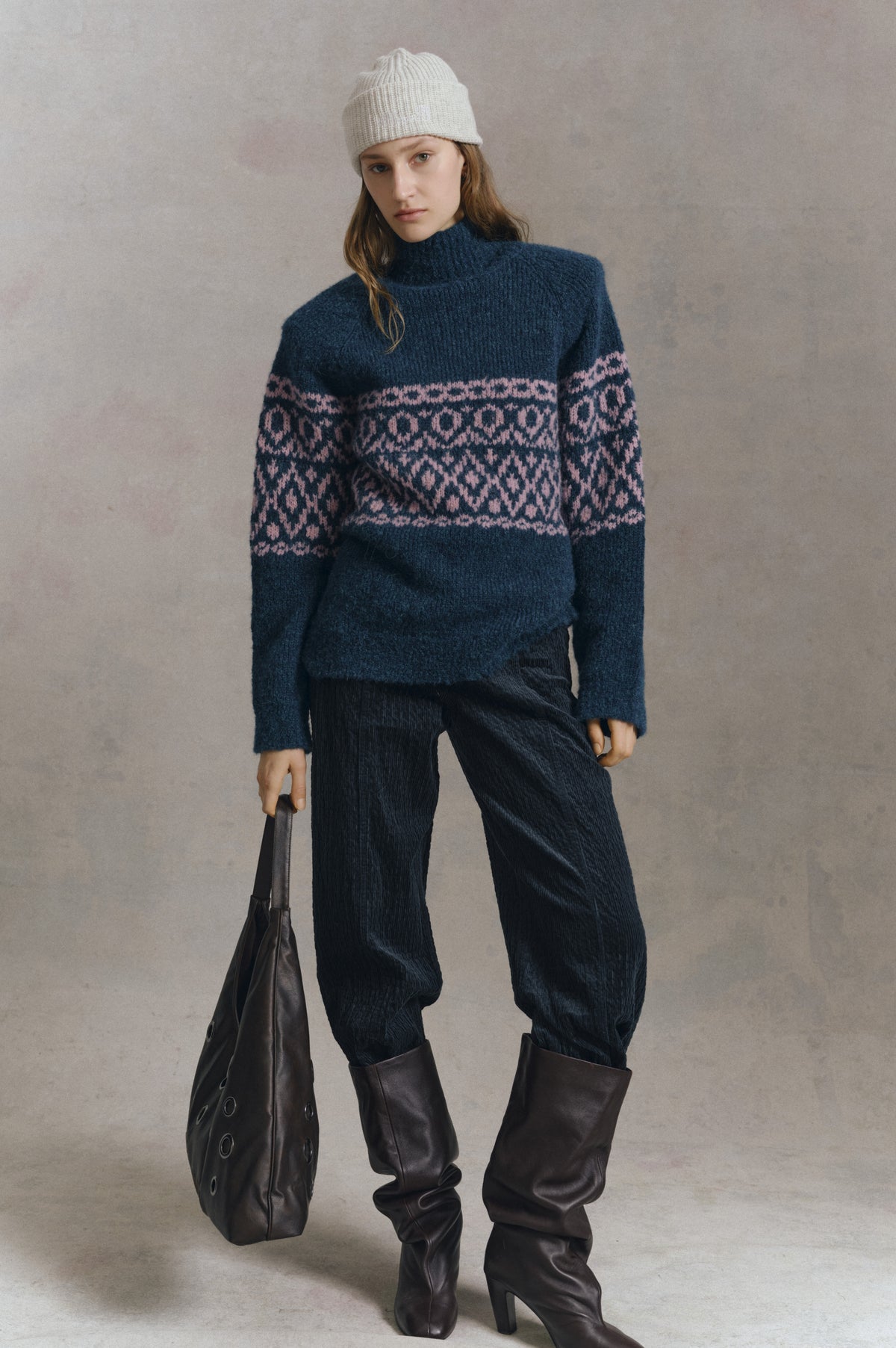 Model wearing a navy high-neck sweater with pink geometric bands, a cream ribbed beanie, dark trousers and tall brown boots, carrying a dark bag.