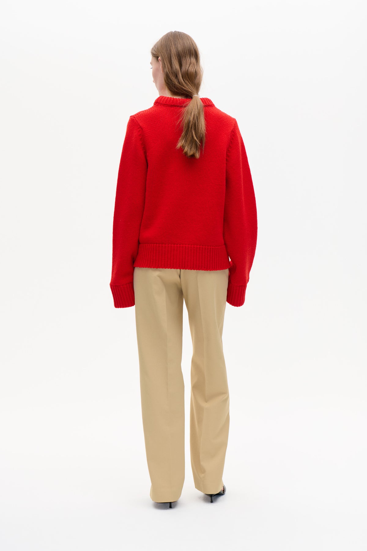 Back view of a model wearing a bright red knitted sweater with a ribbed collar, cuffs and hem, paired with beige trousers.