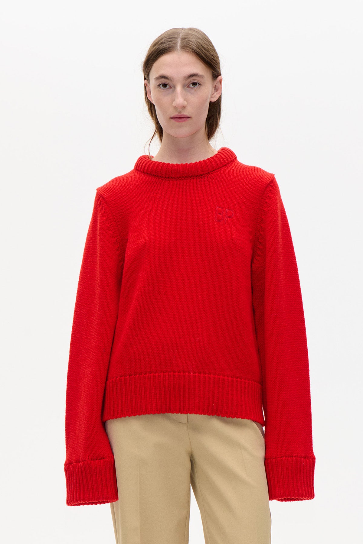 Front view of a bright red knitted sweater with a ribbed crew neckline, wide cuffs and hem, worn with beige trousers.
